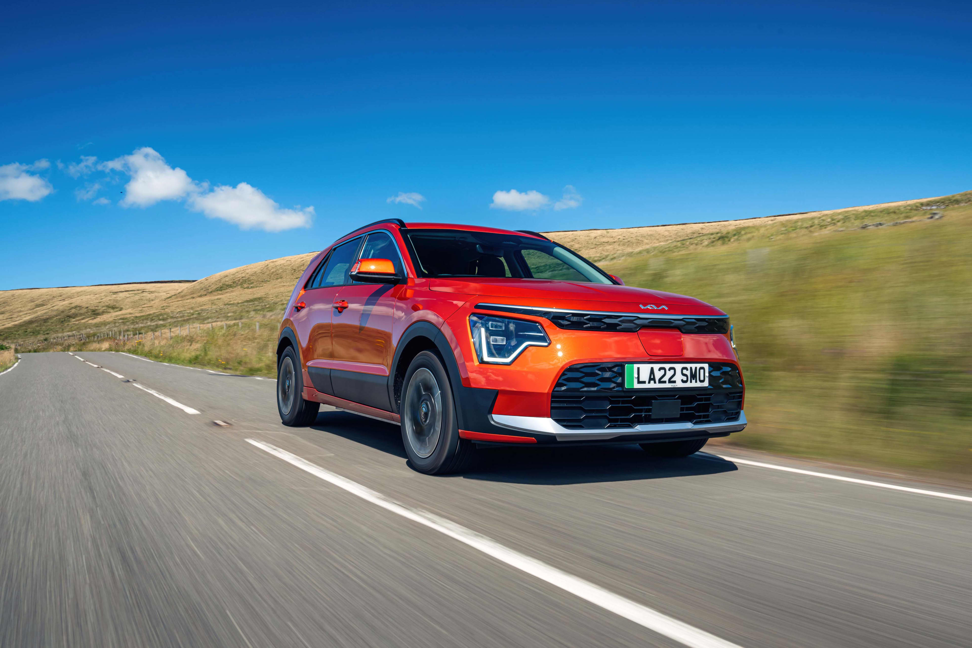 A red/orange Kia Niro EV, model 2022- current, drives along a bitumen road with grassed hills blurred in the background.