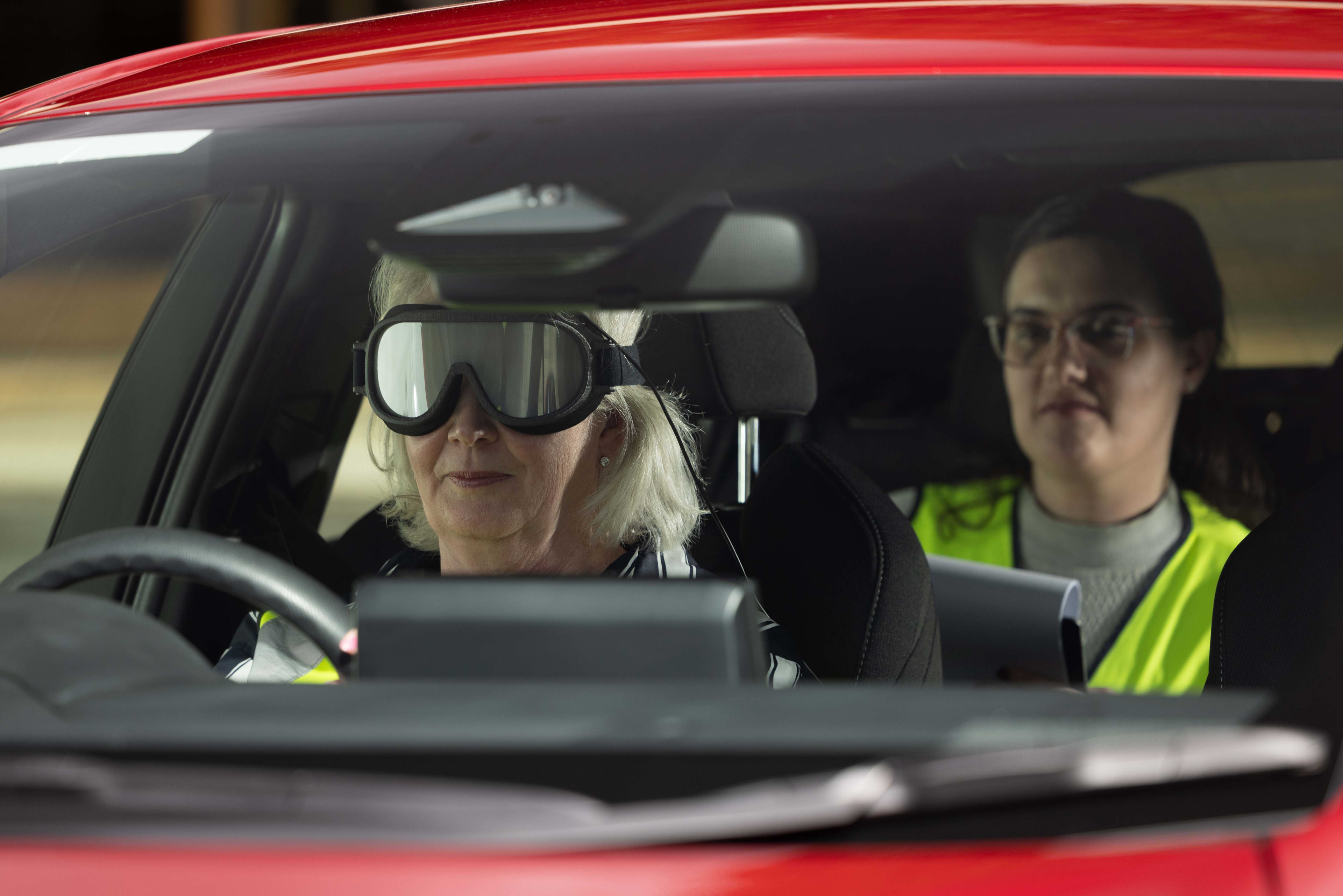 A female driver sits in the front of a red car, wearing eye-tracking goggles. A female in a high vis vest sits in the back holding a clipboard.