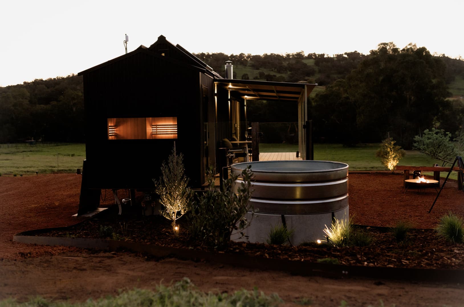 A dusk image of a black chalet with a window showing the sauna lit up and a metal plunge pool sitting beside.