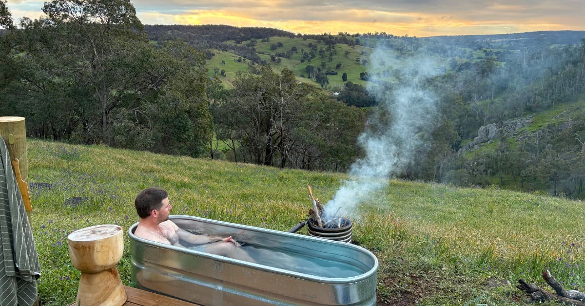 An image of a metal tub filled with water with a male sitting inside overlooking the rolling farmland. There's a heated coil next to the metal tub filled with wood with smoke rising from it.