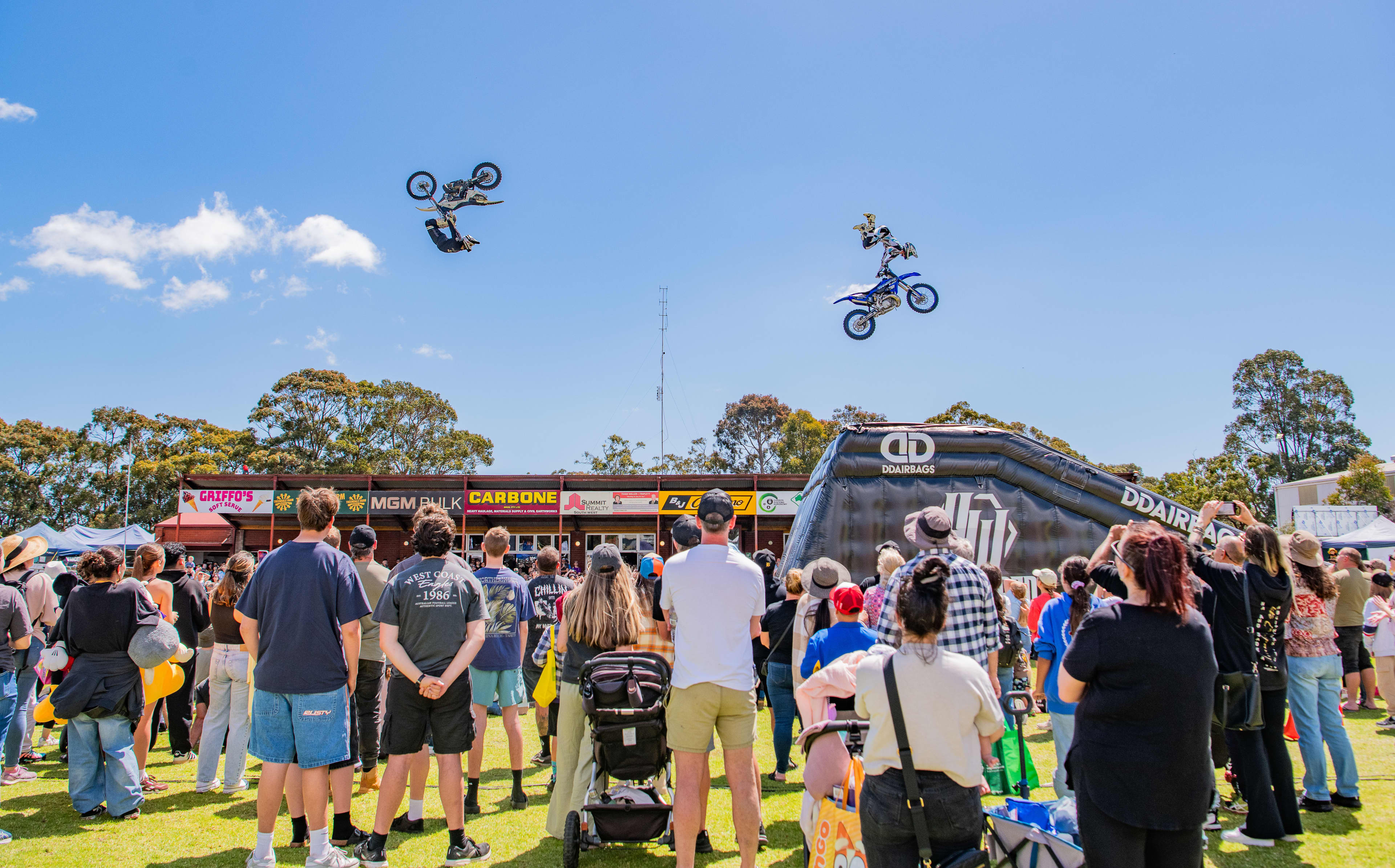 Two motorbikes flying through the air with riders performing tricks, while a crowd gathers to watch.