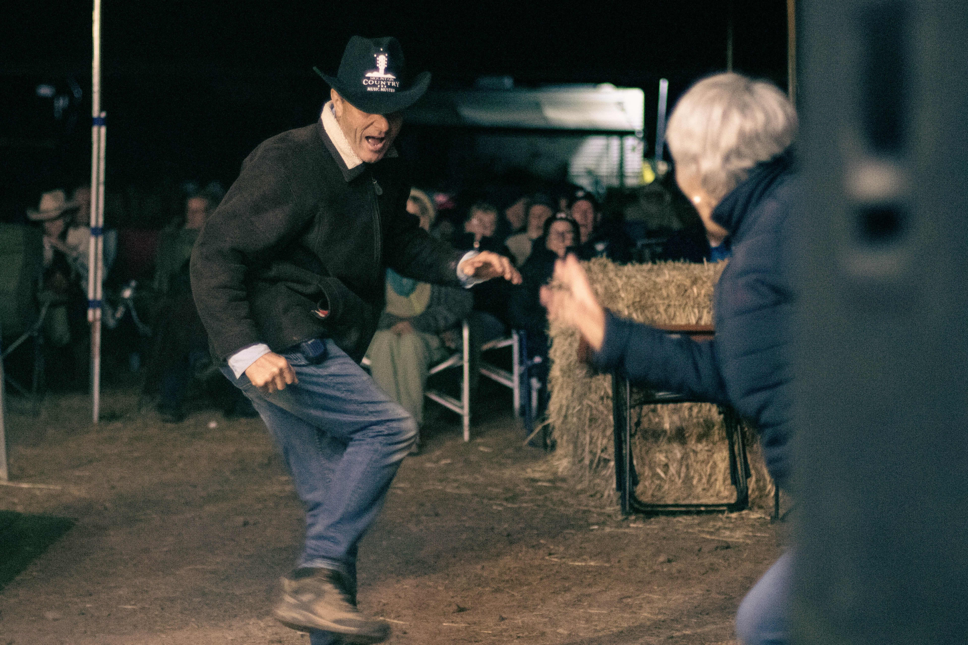 A man wearing a black cowboy hat that reads "country music muster' dancing with a partner with festival goers watching in the background.