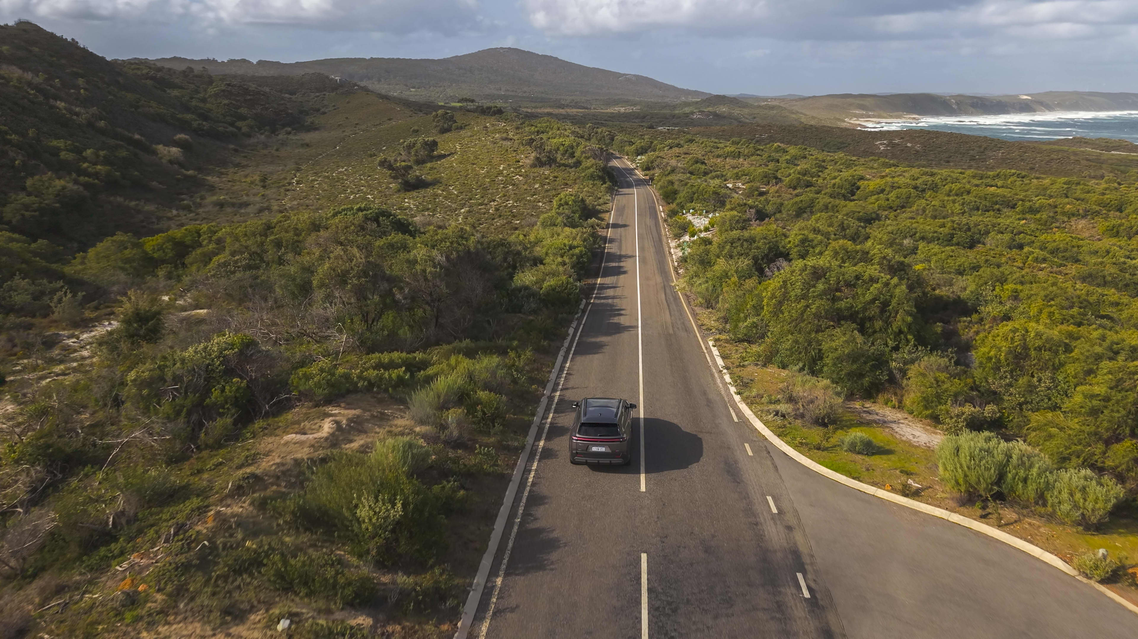 An EV drives down a road in WA's South West, surrounded by greenery and with ocean in the distance.