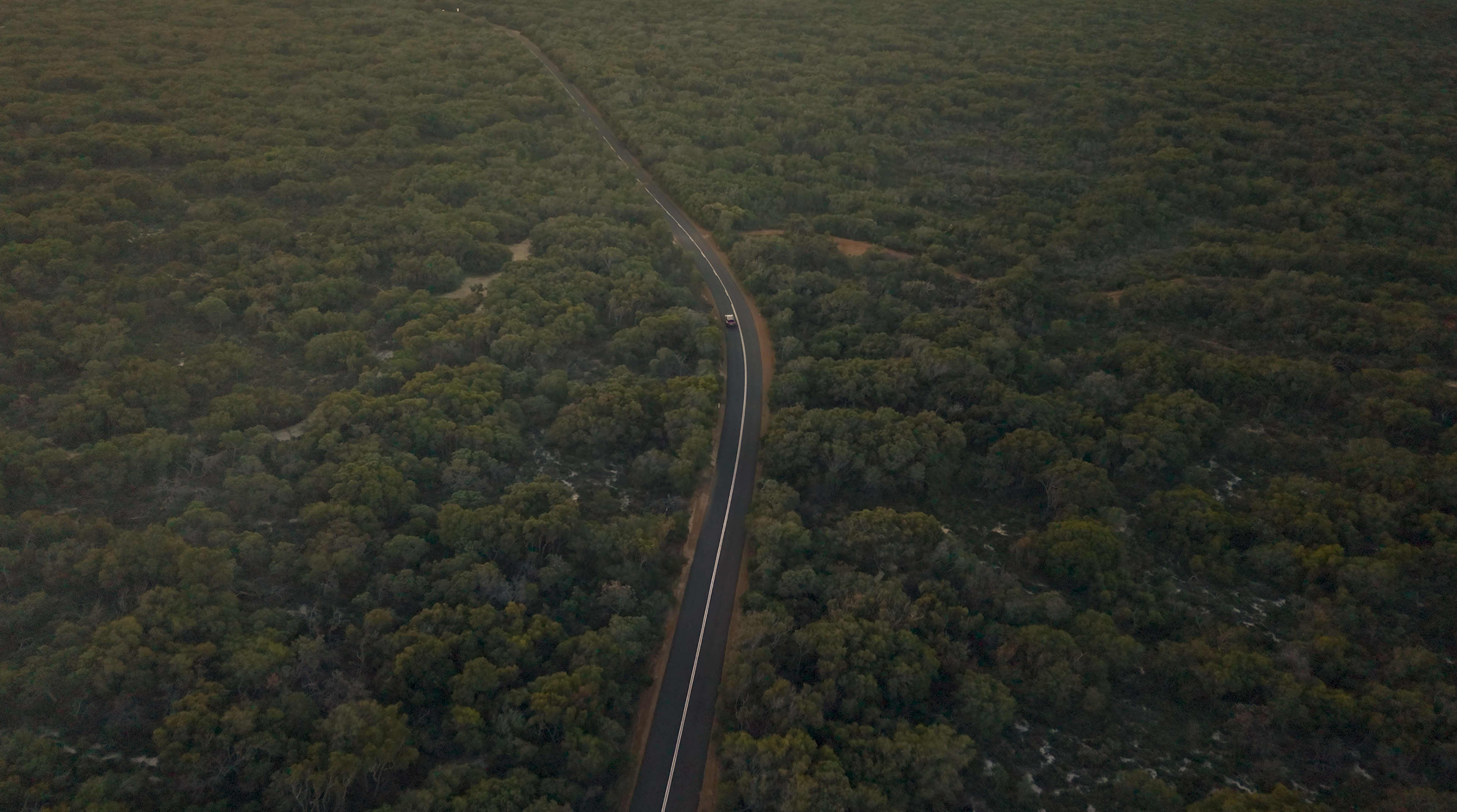 A distant aerial shot of a road cutting through bushland, with an EV just visible driving along.