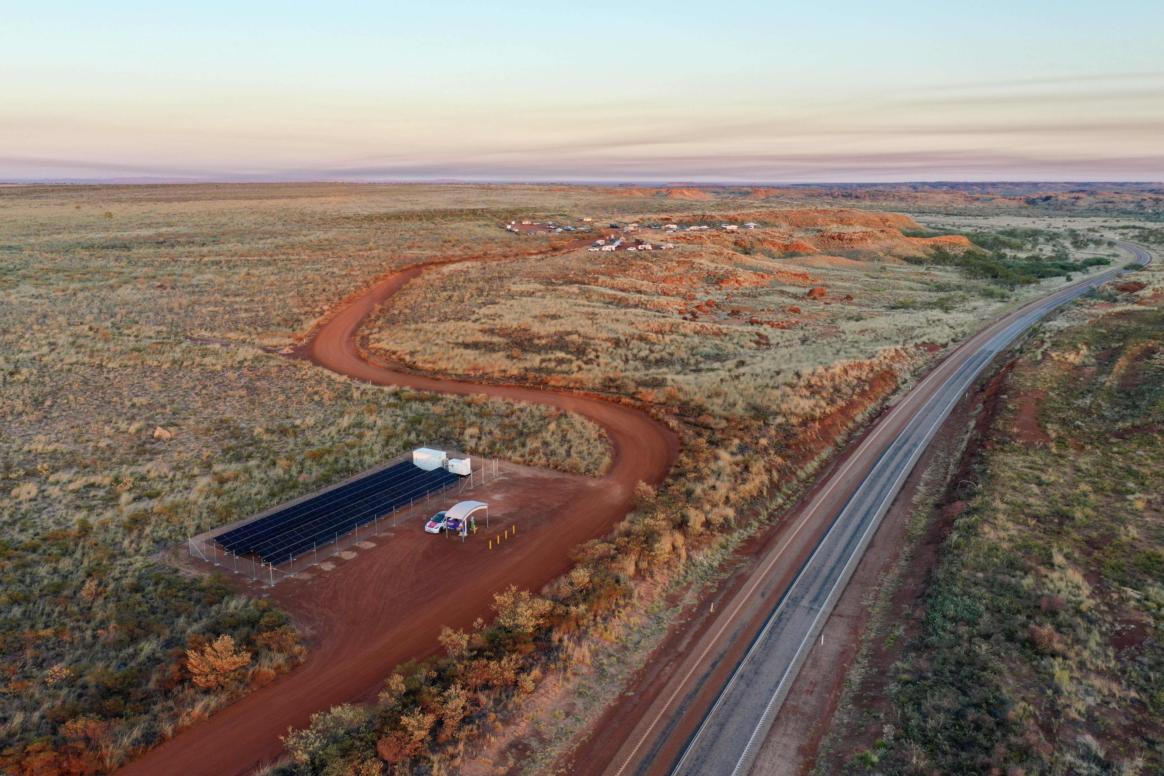 An aerial shot of a remote WA landscape showing a road running through and a cleared area with an EV charger, with a car parked and solar panels.