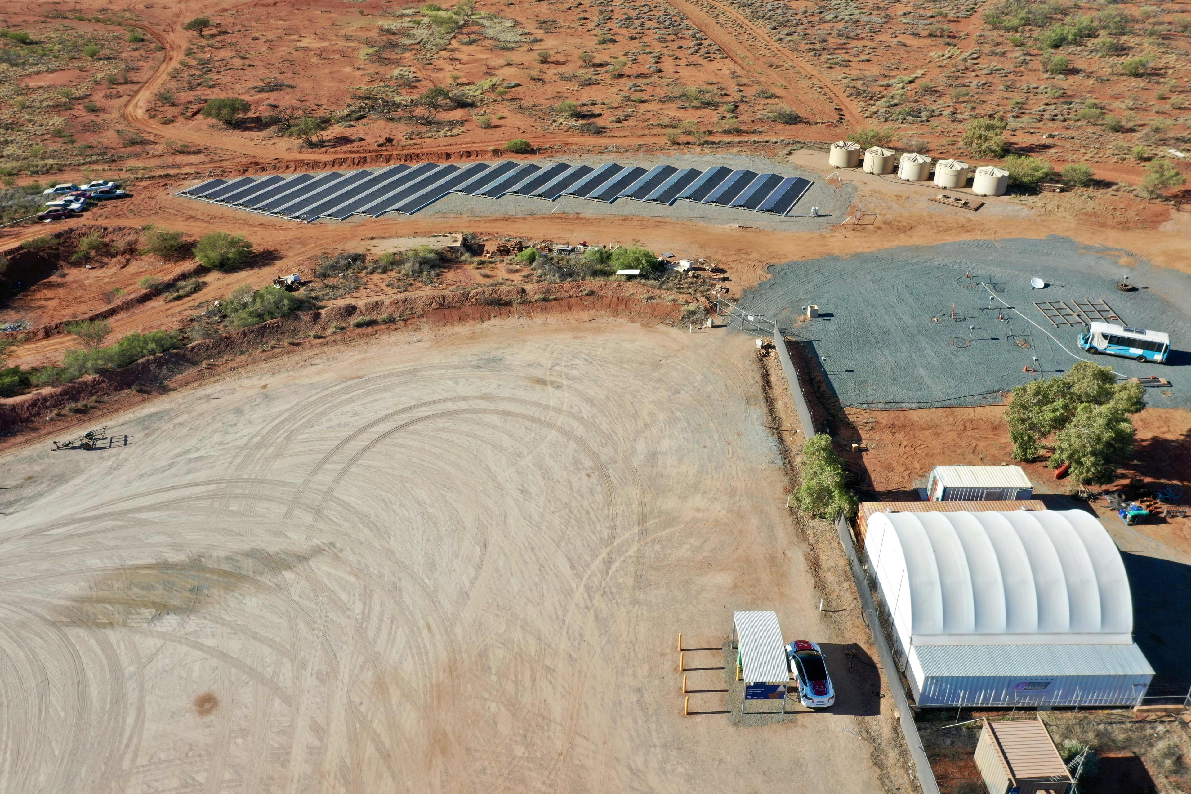 An aerial shot of a cleared area with an EV charger and solar panels, surrounded by red dirt and scrub.