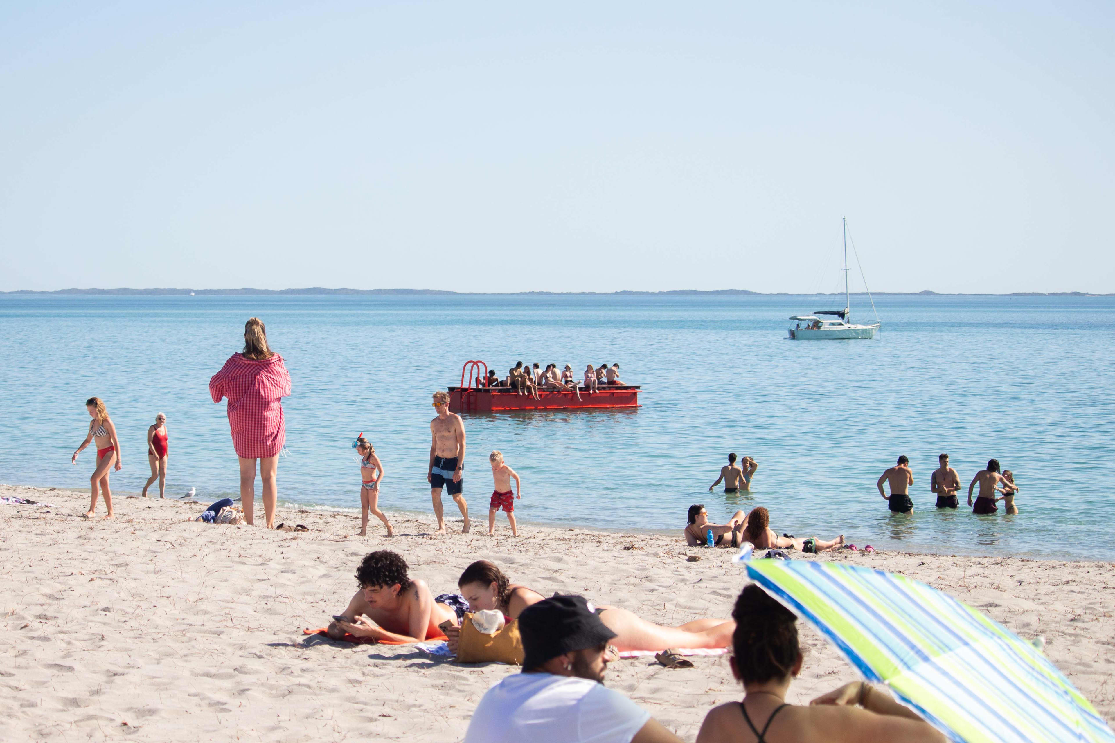People lie or walk on the sandy beach on a sunny day, with a red pontoon full of swimmers in the background.