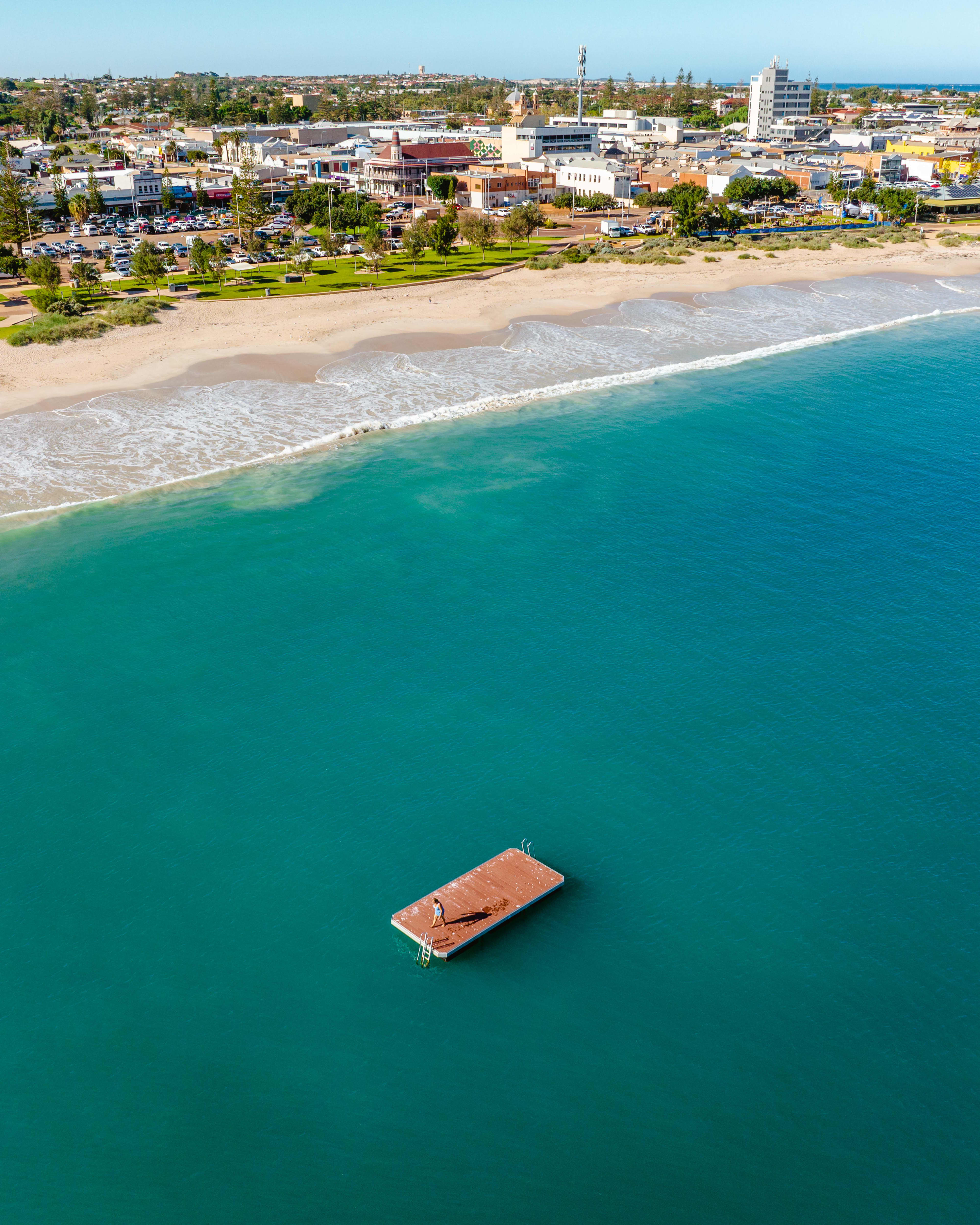 An aerial view of a red pontoon on a calm blue ocean, with the beach in the background and the town further back.