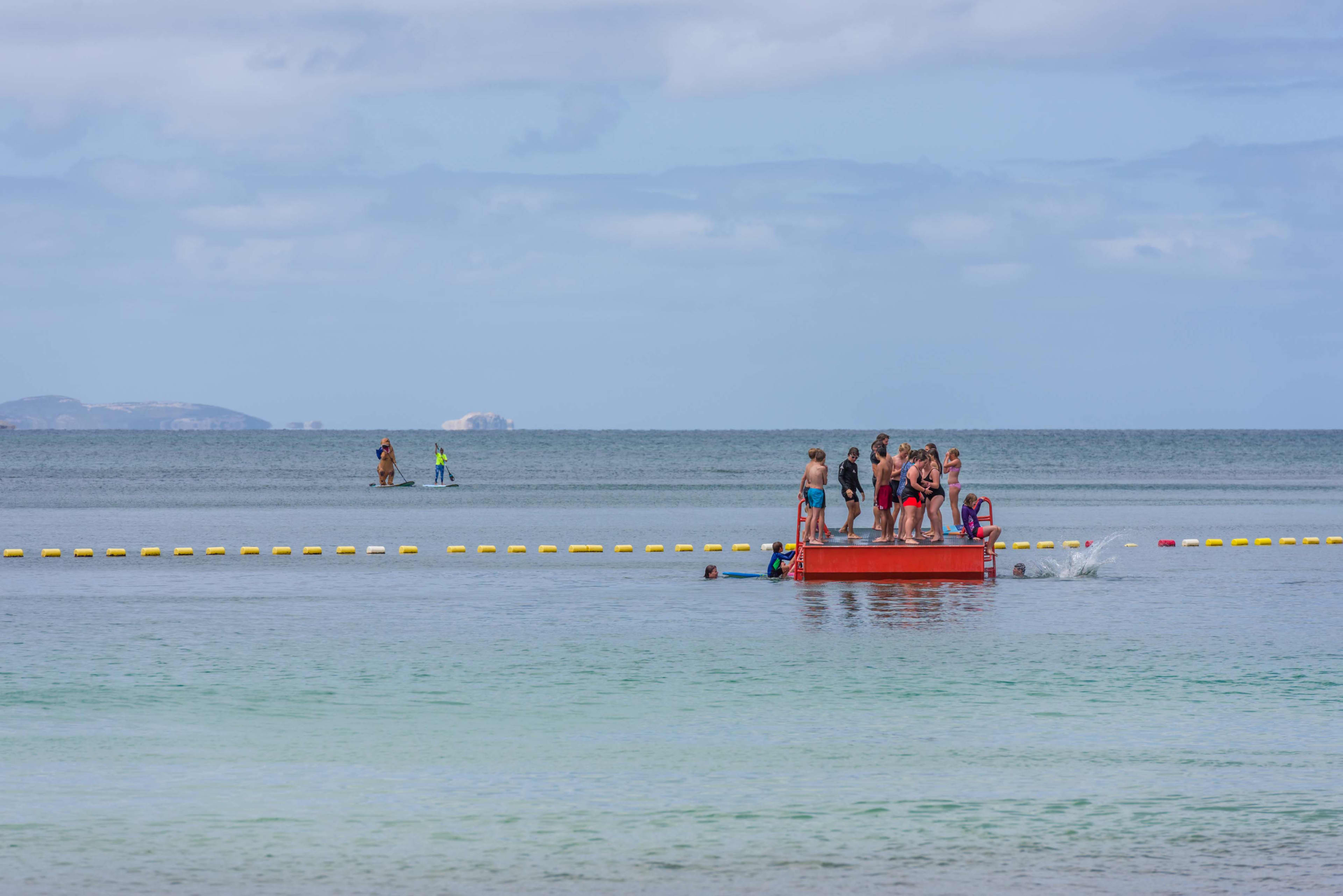 A crowd of people in bathers standing on and jumping off a red pontoon in the blue ocean, with yellow buoys in a line behind them.