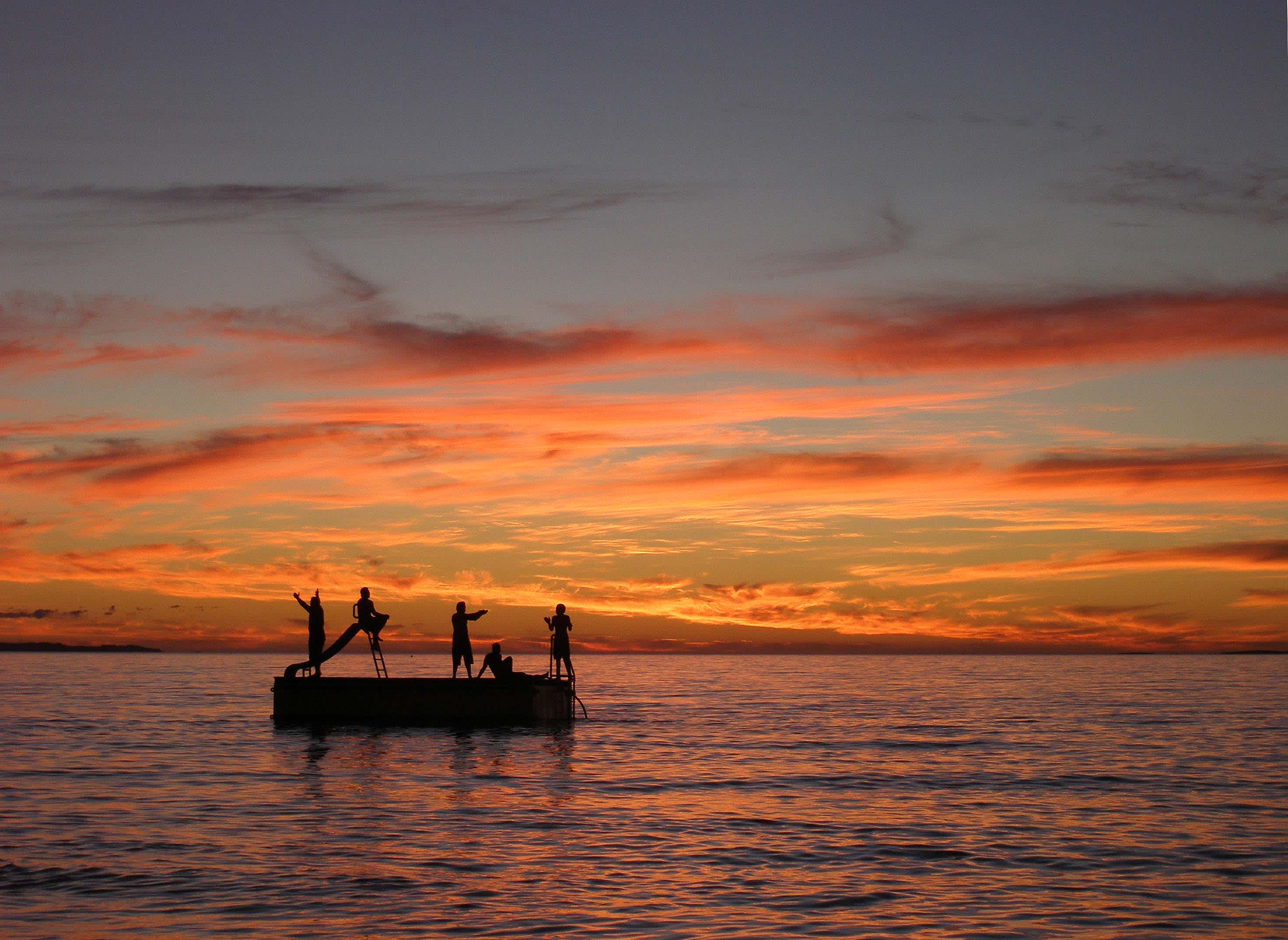 People silhouetted against the sunset, standing on and jumping off a pontoon in the ocean.