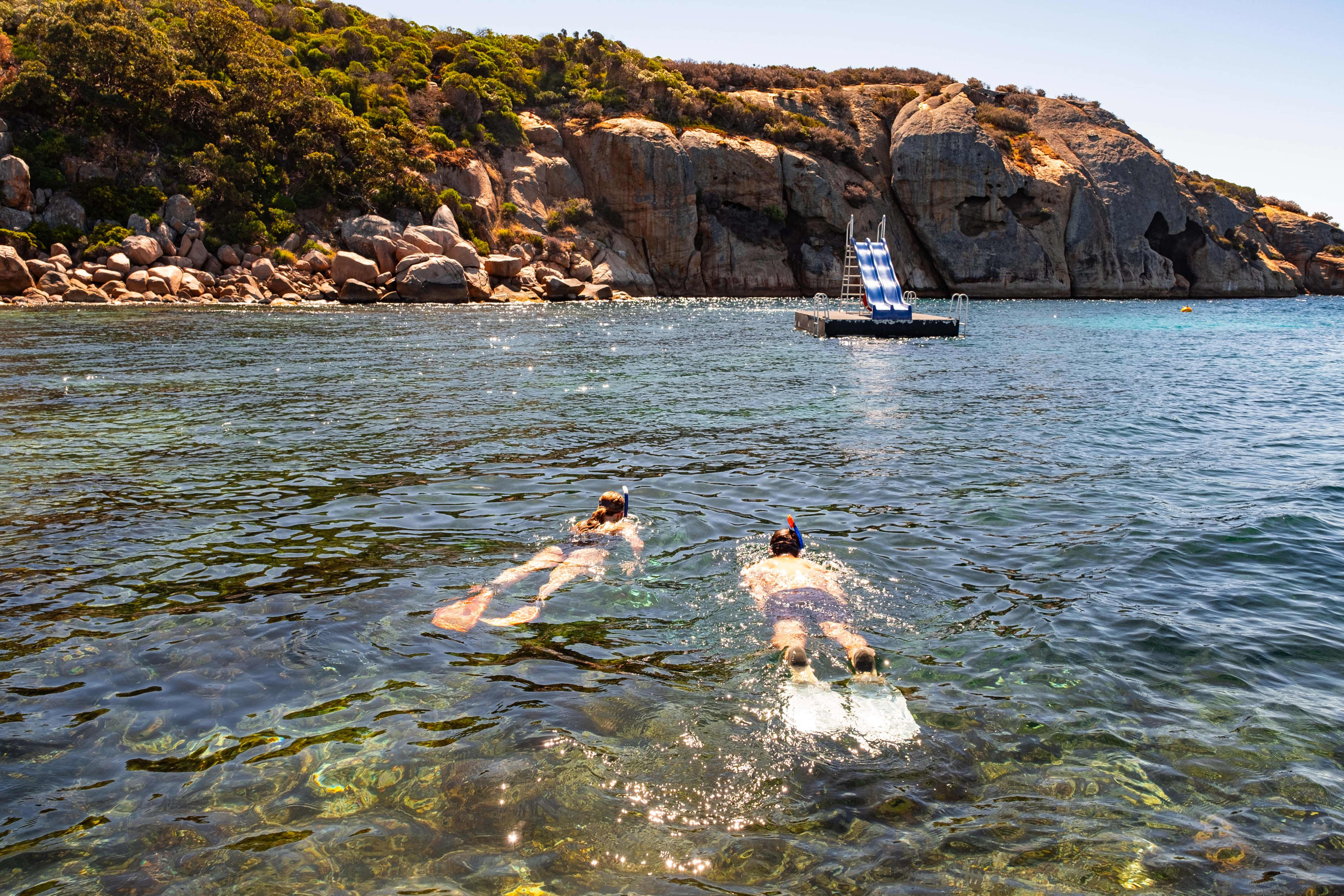 A man and a woman snorkel over a reef towards a pontoon with a blue slide, with the rocky cliff of an island in the background.