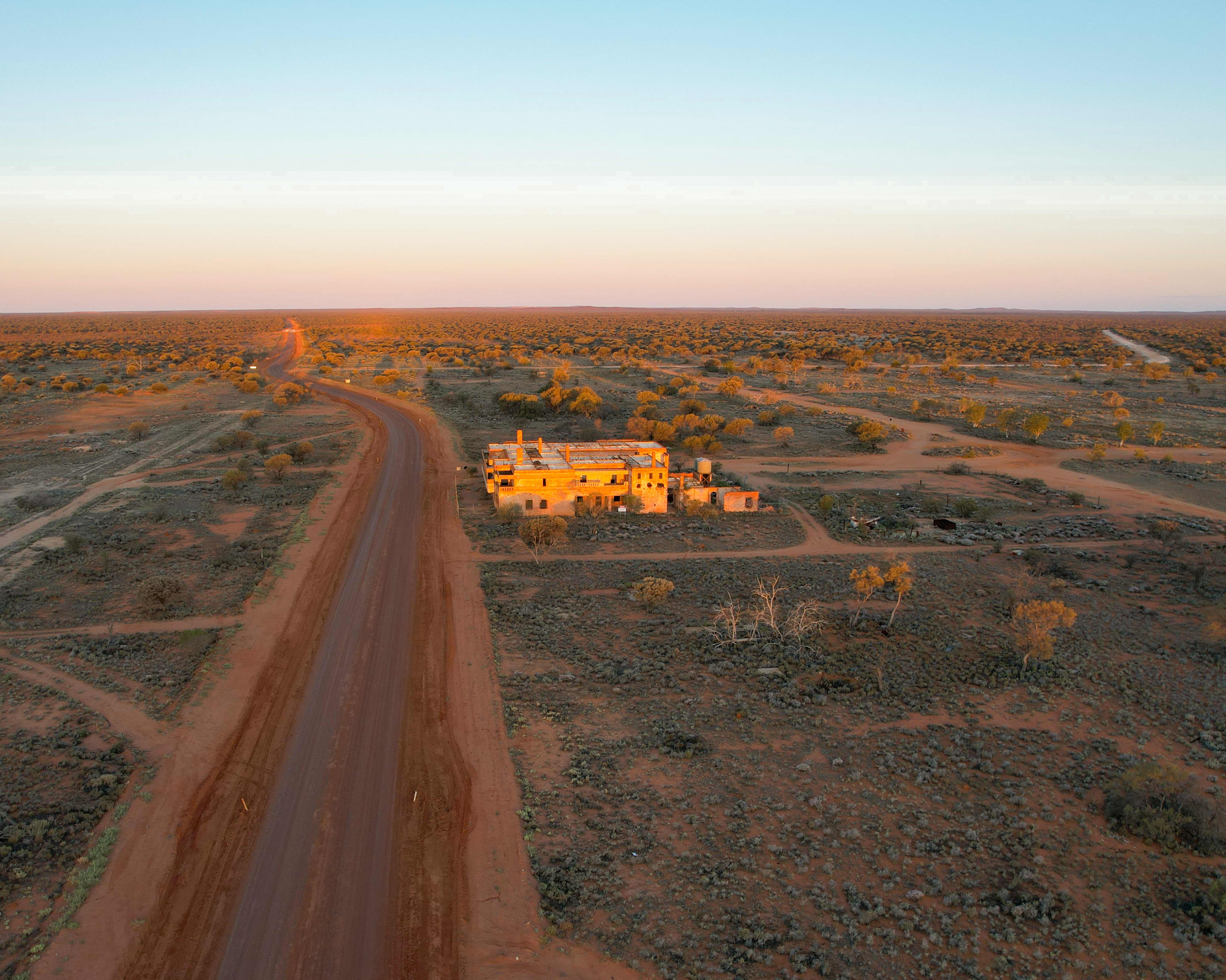 An aerial, wide-angle shot captures a remote building situated in a vast, desert landscape under a clear sky during sunset.