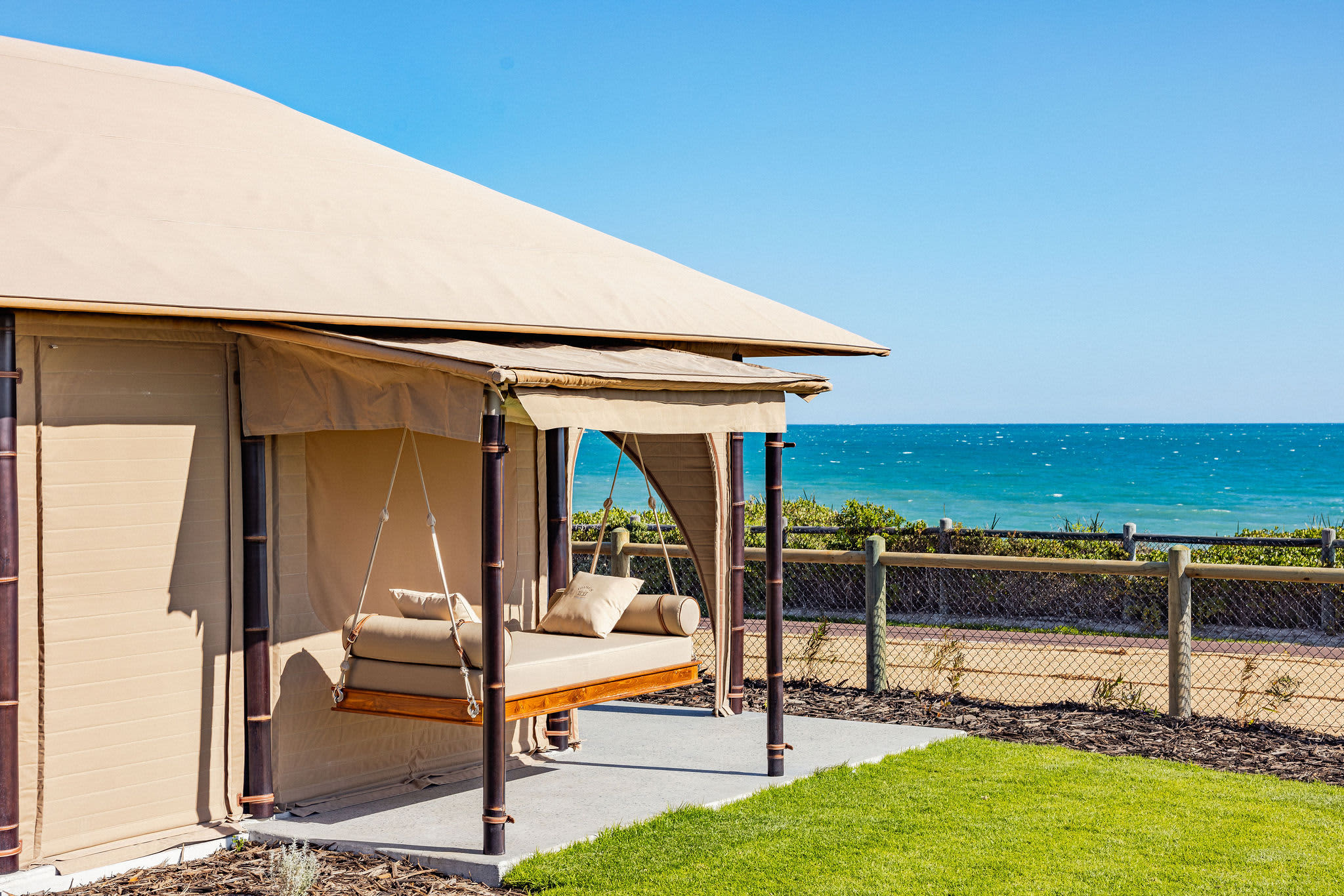 A coastal luxury cabana featuring a swinging daybed sits on a green lawn overlooking a vibrant turquoise ocean.