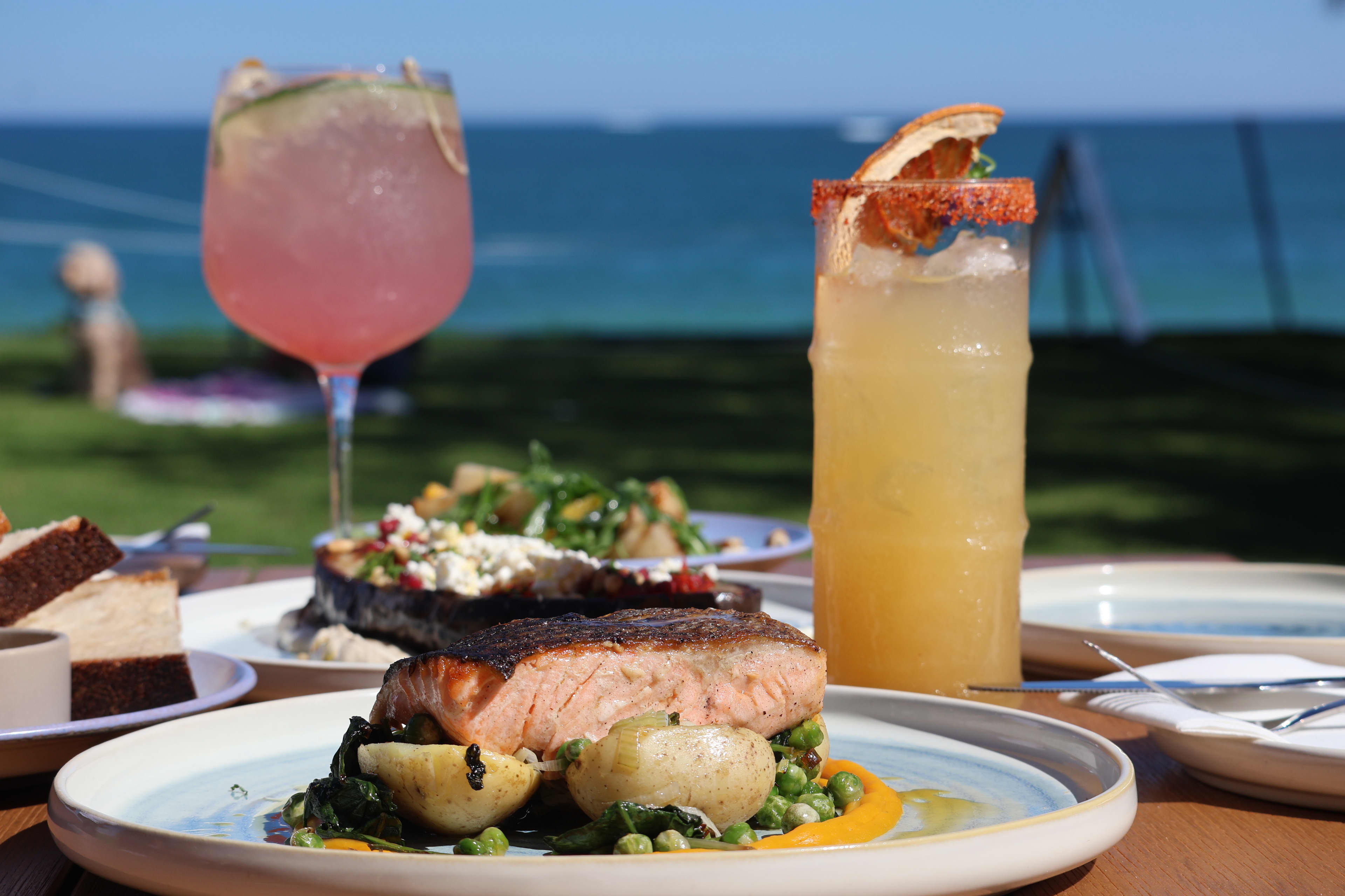 A vibrant beachside dining layout with seared salmon fillet accompanied by small potatoes, green peas and greens.