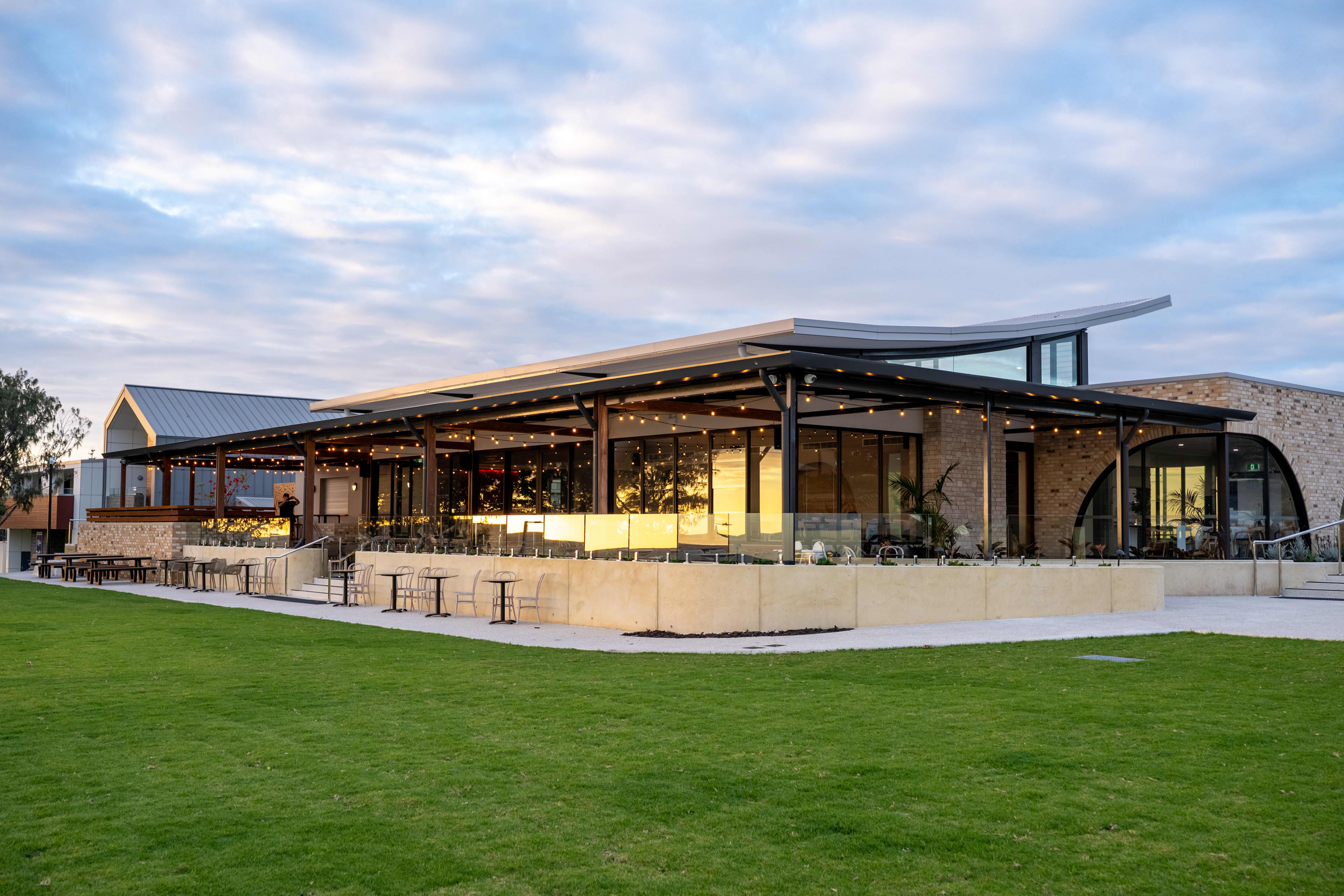 A modern, single story commercial venue featuring an extensive outdoor dining patio shaded by a dark architectural awning adorned with warm fairy lights.