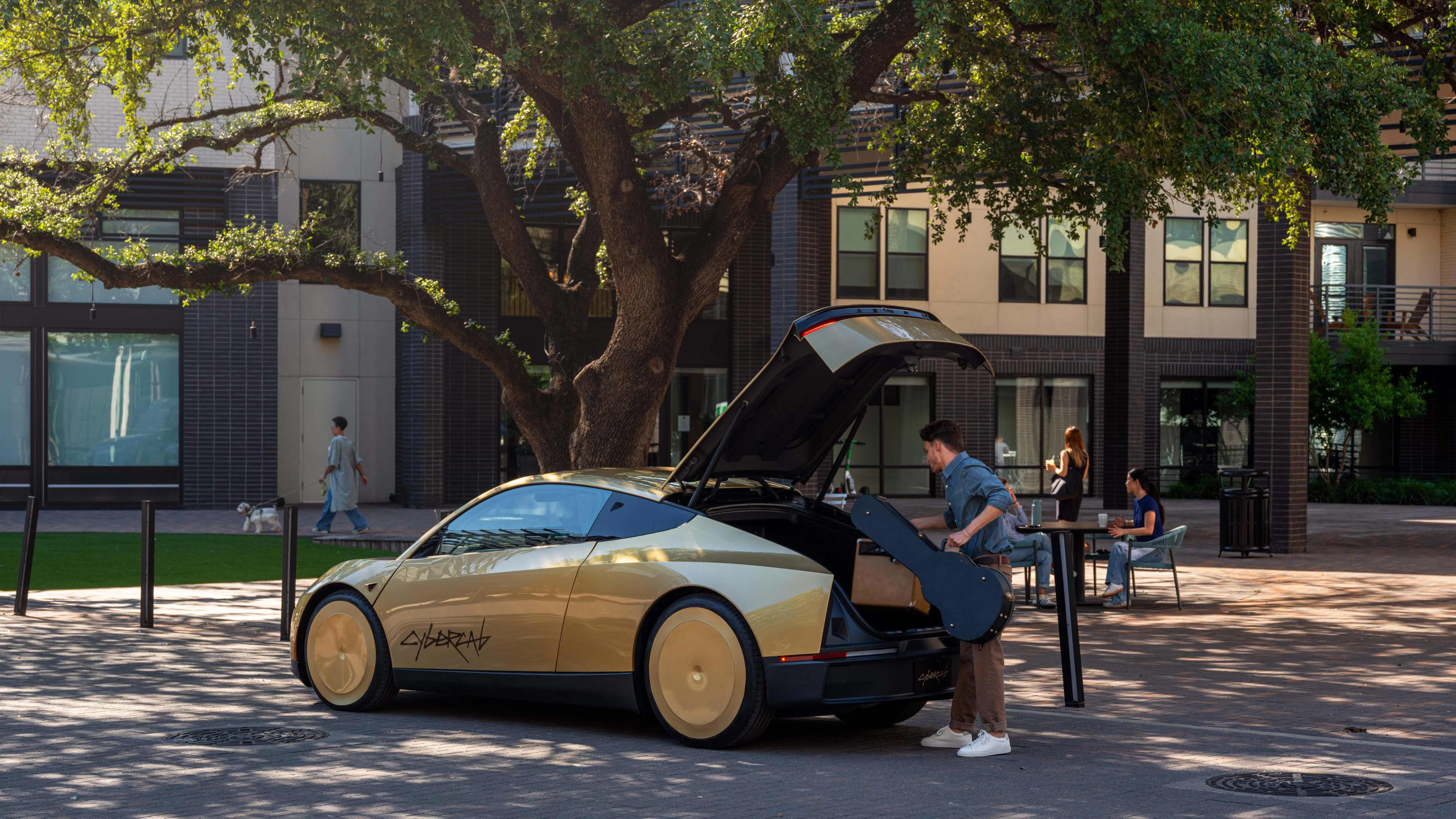 A man puts a guitar case into the boot of a golden car with Cybercab written on the door, parked under a tree outside some buildings.