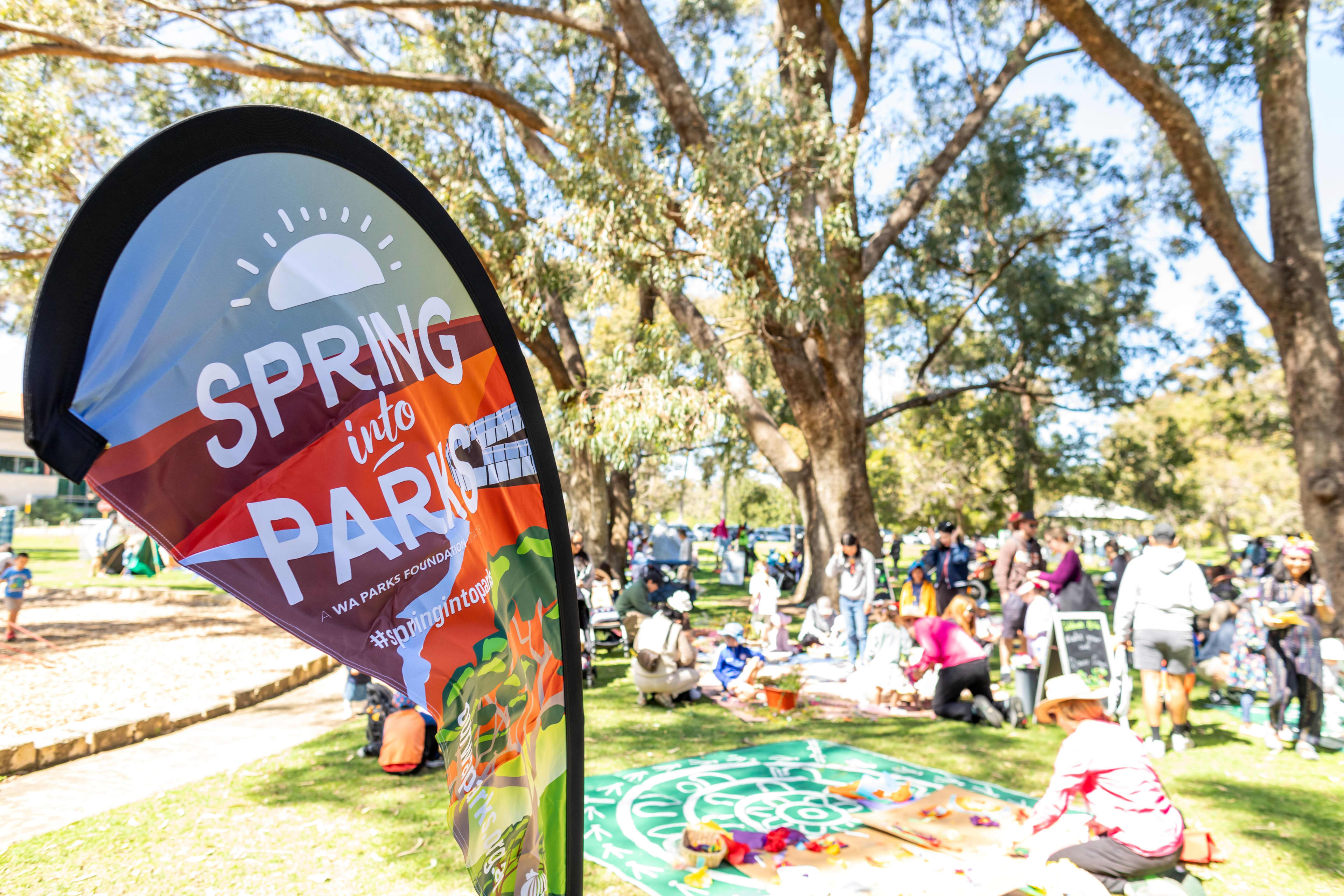 A vibrant teardrop-shaped outdoor banner in the foreground, with the colourful text 'Spring into Parks' printed prominently on it.