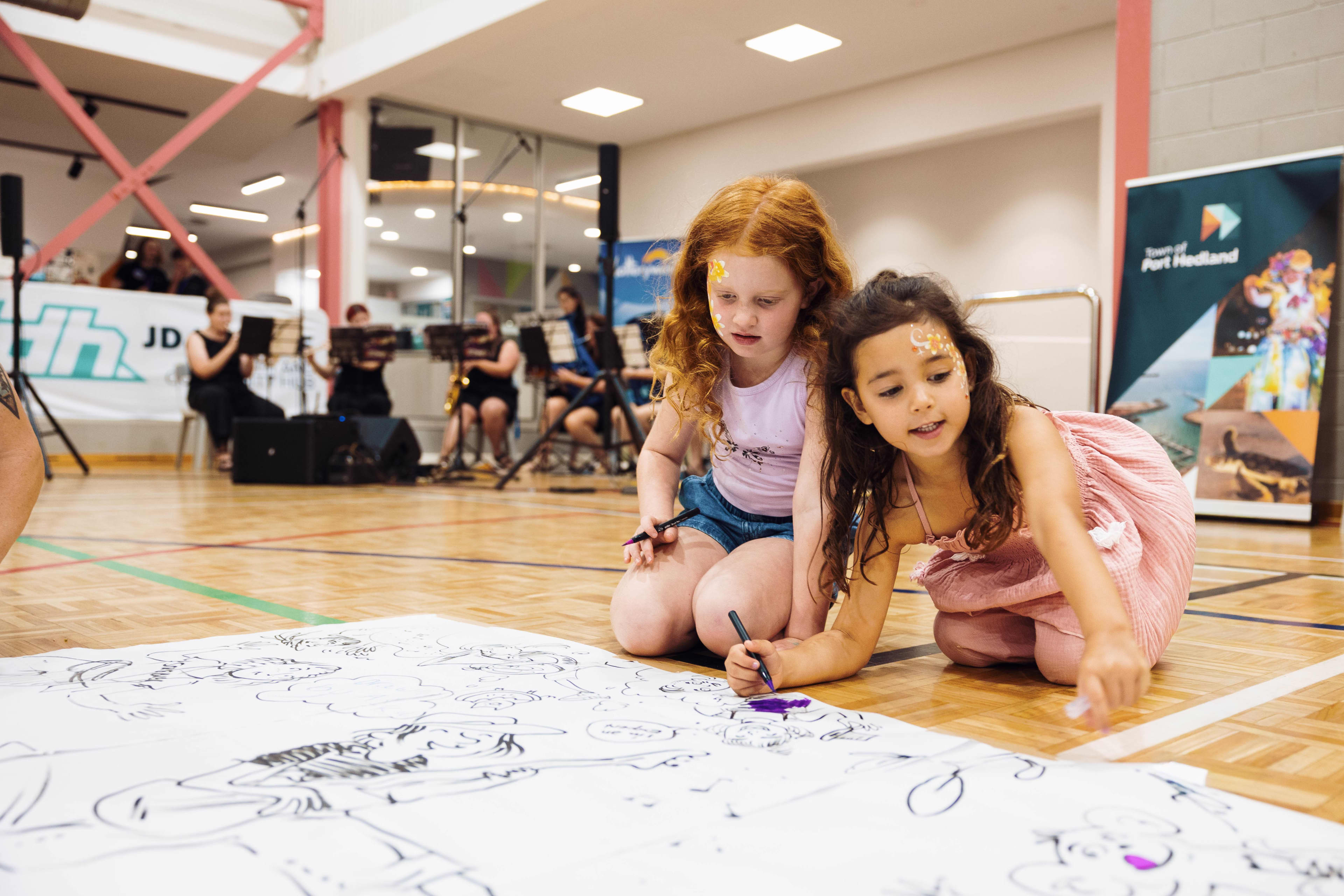 Two young girls with face paint kneel on a wooden floor inside a community centre, colouring on a large sheet of paper spread out before them.