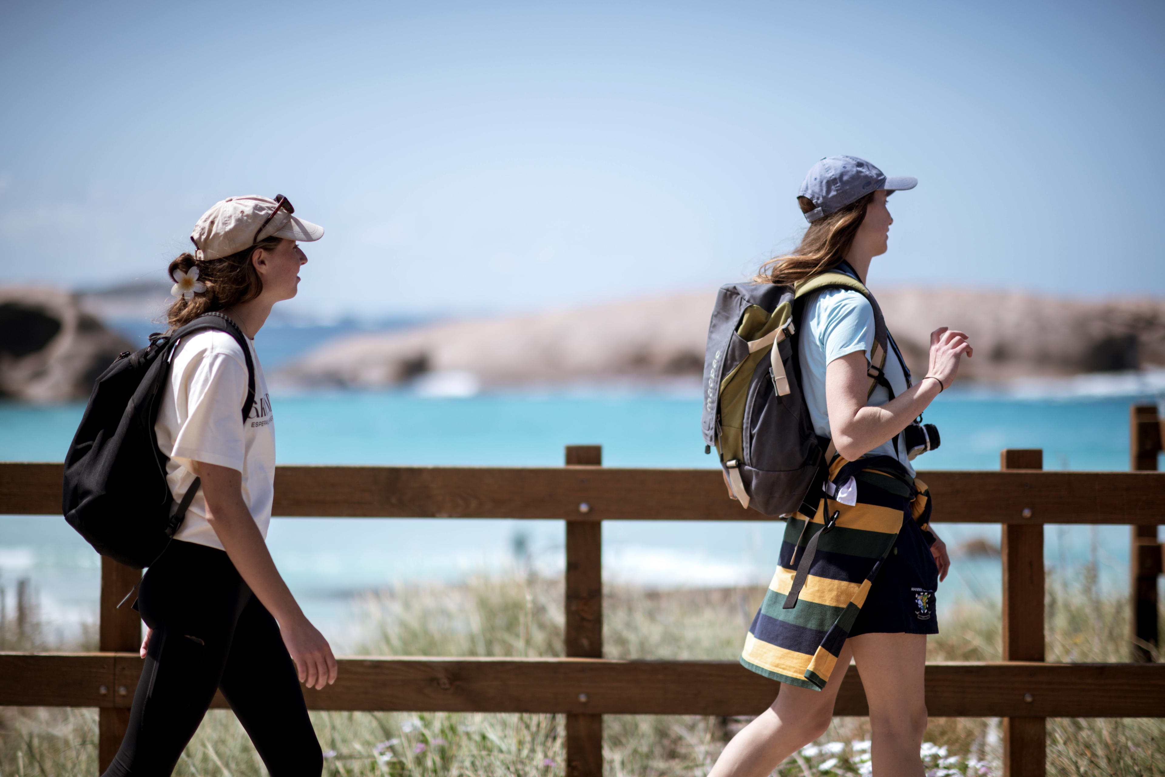 Two hikers walking along a wooden boardwalk overlooking a bright blue coastline.