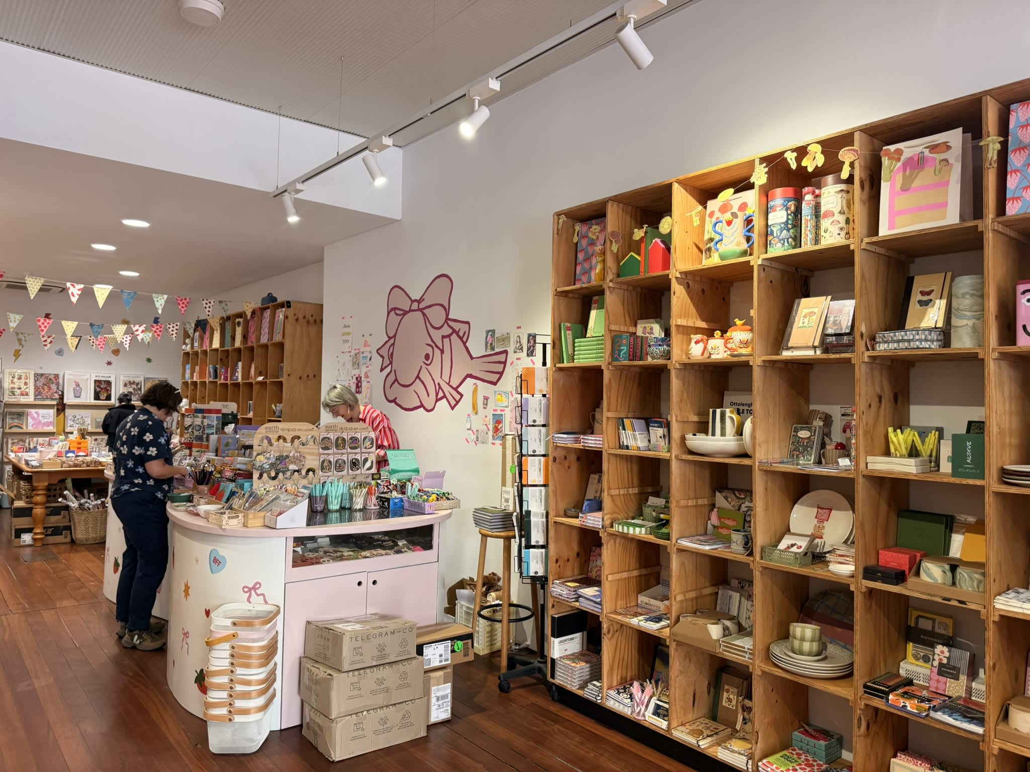 Colourful stationery shop with wooden pigeon holes full of pens and notebooks and a counter in the centre of store.