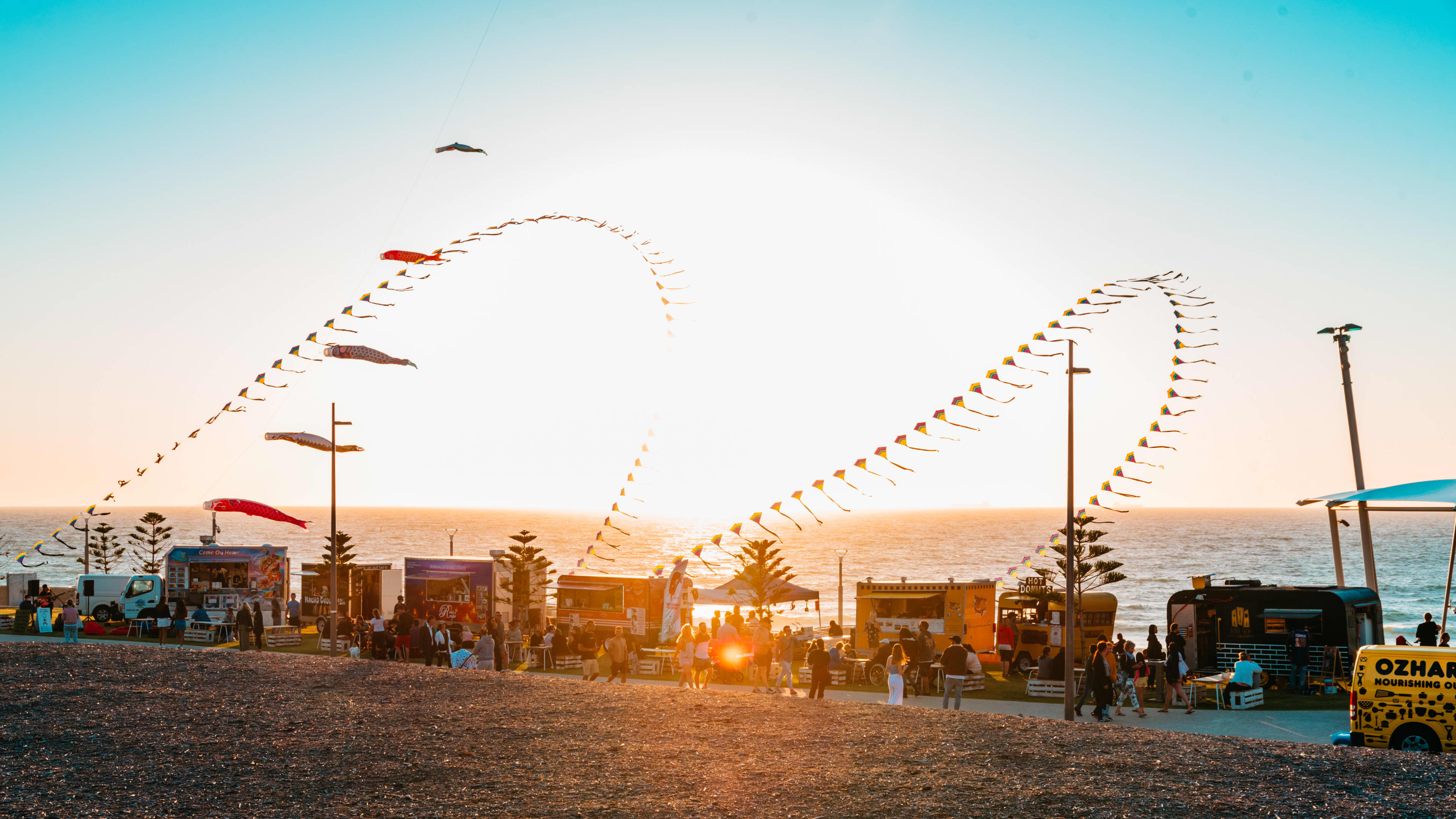 An image at dusk with a line of food trucks sitting in front of the ocean front, with large decorational kites flying in the wind.