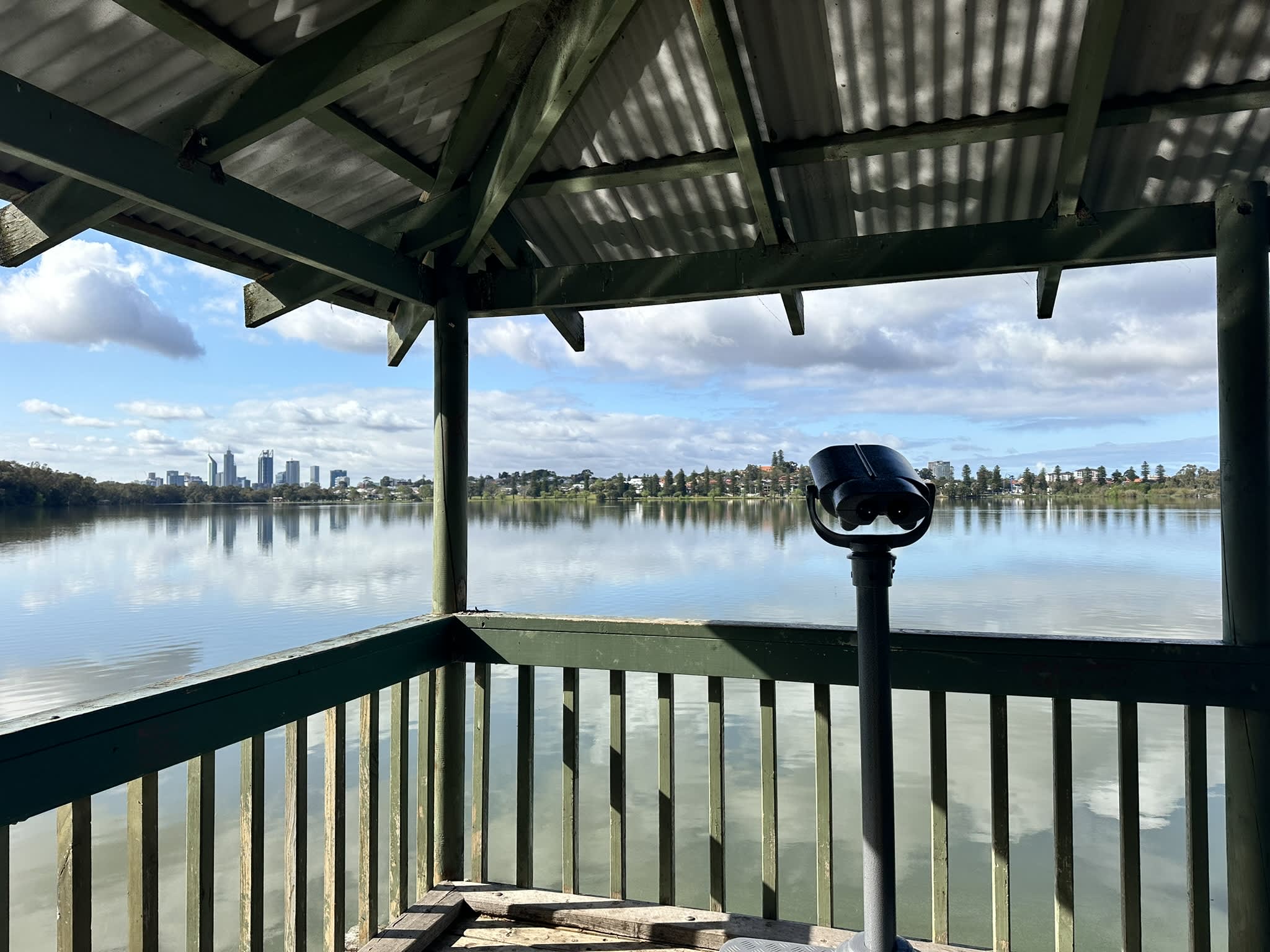 Binoculars inside a gazebo overlooking a lake surrounding by trees with city skyline in background.