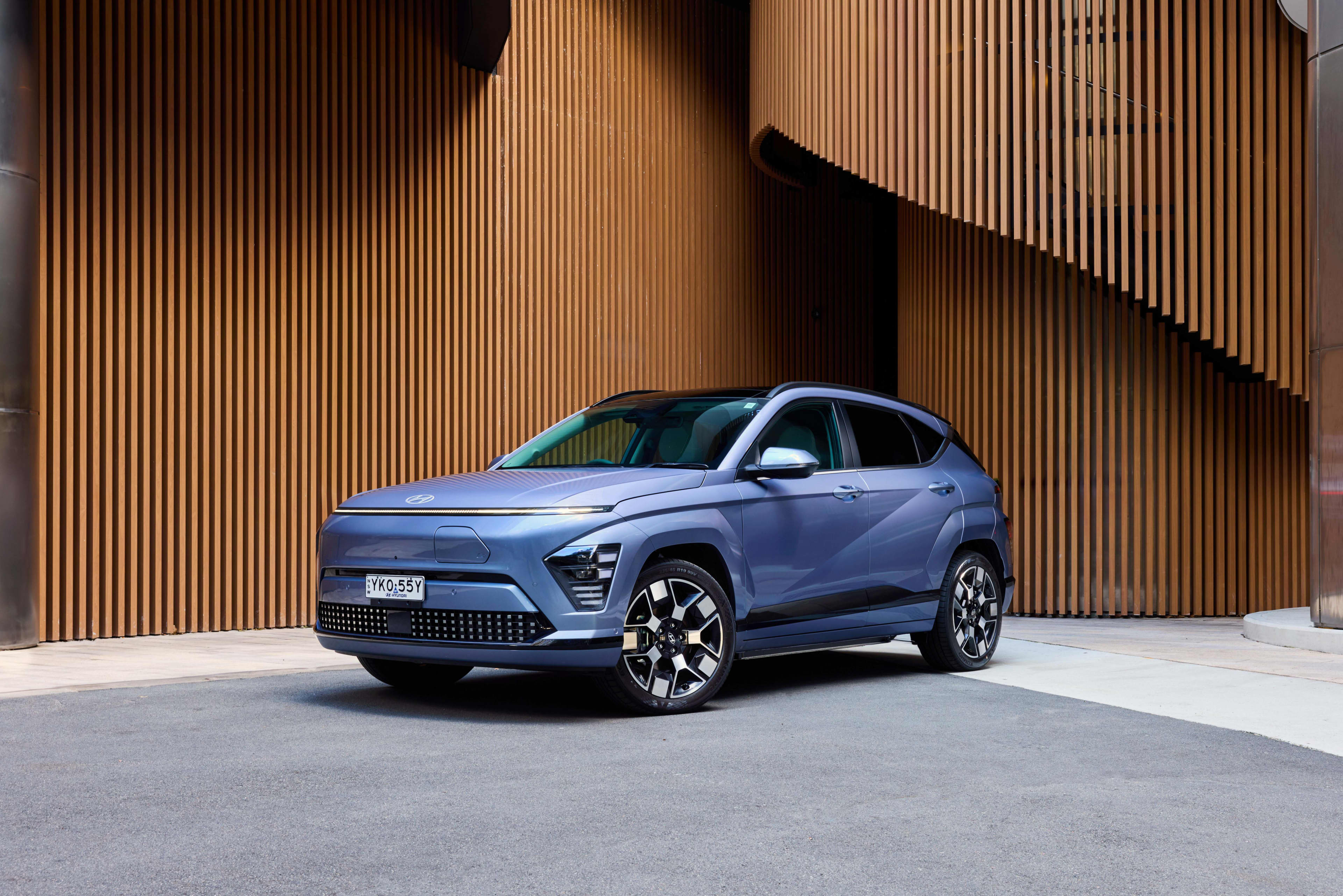 A blue Hyundai Kona Electric car parked outside a building with wooden panel detailing.