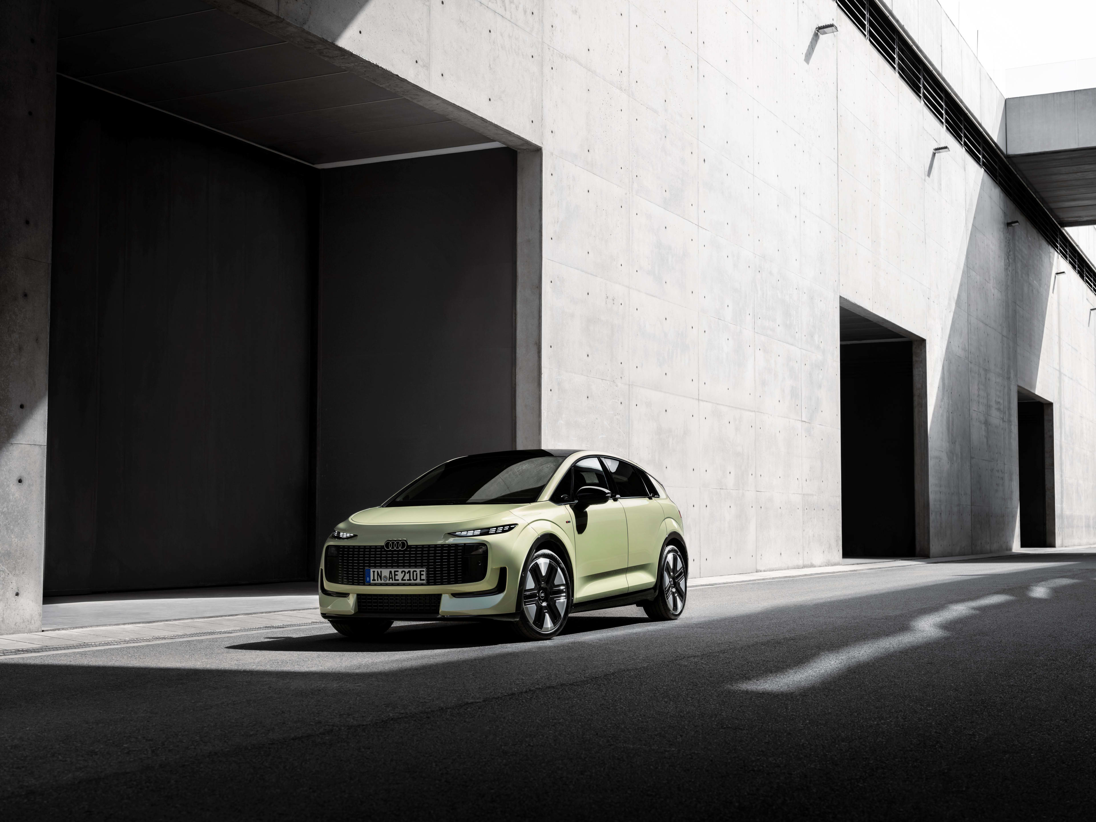 A light green car driving along a city street with a concrete wall behind.