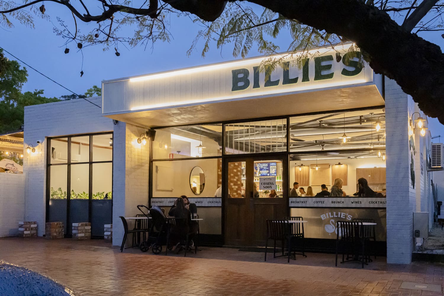 A brightly lit, modern cafe storefront named "Billies" at dusk.