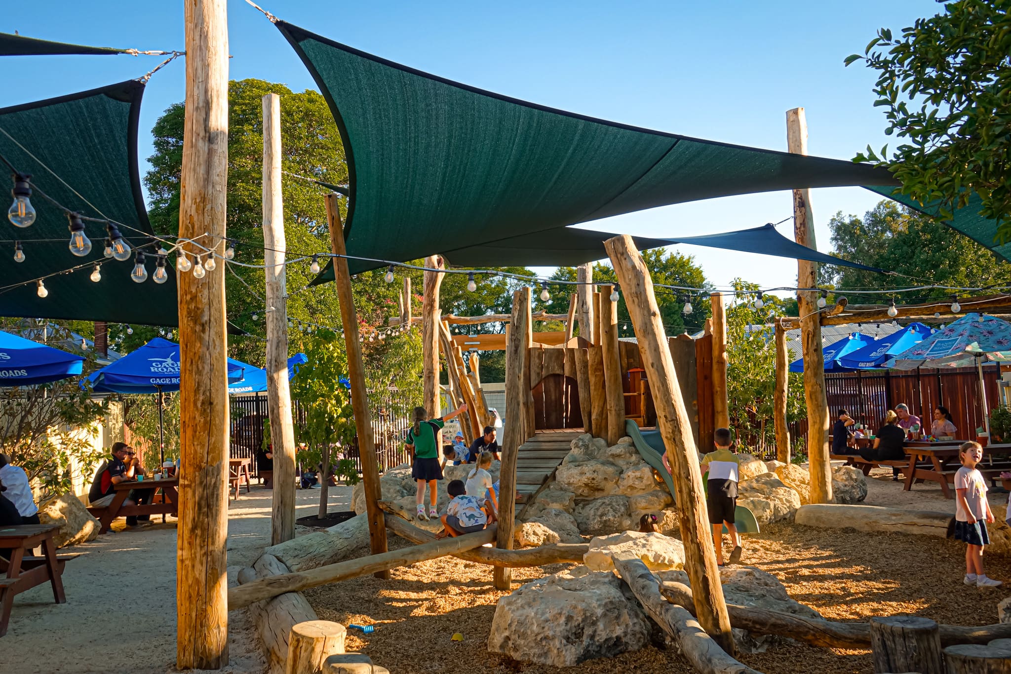 An outdoor nature-based playground at a family-friendly venue is filled with children playing under large shade sails.