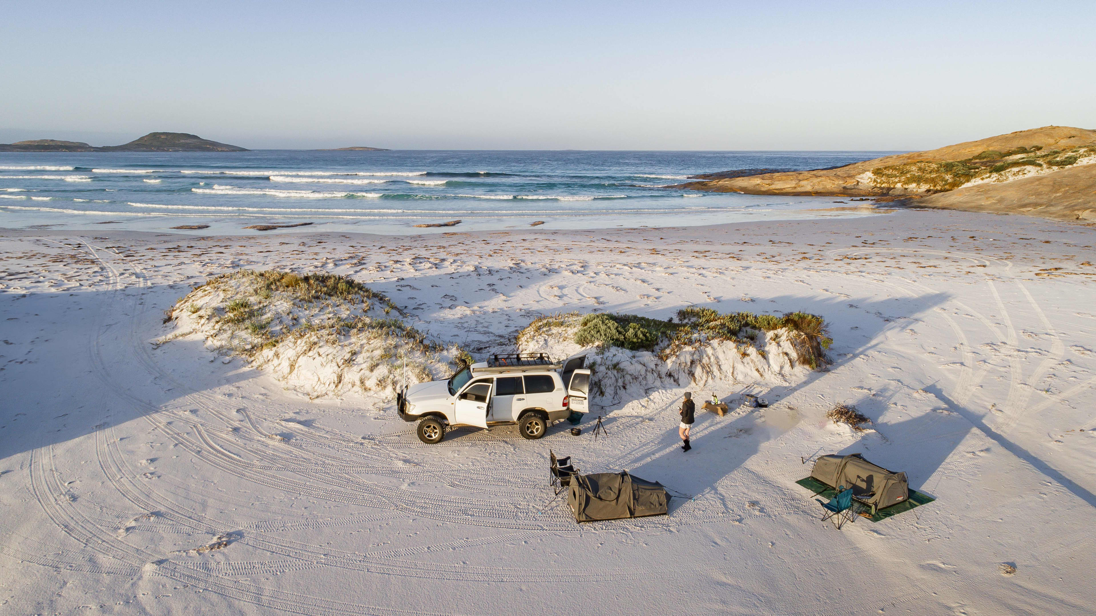 A white 4WD and two khaki swags set out on a beach with ocean in the background.