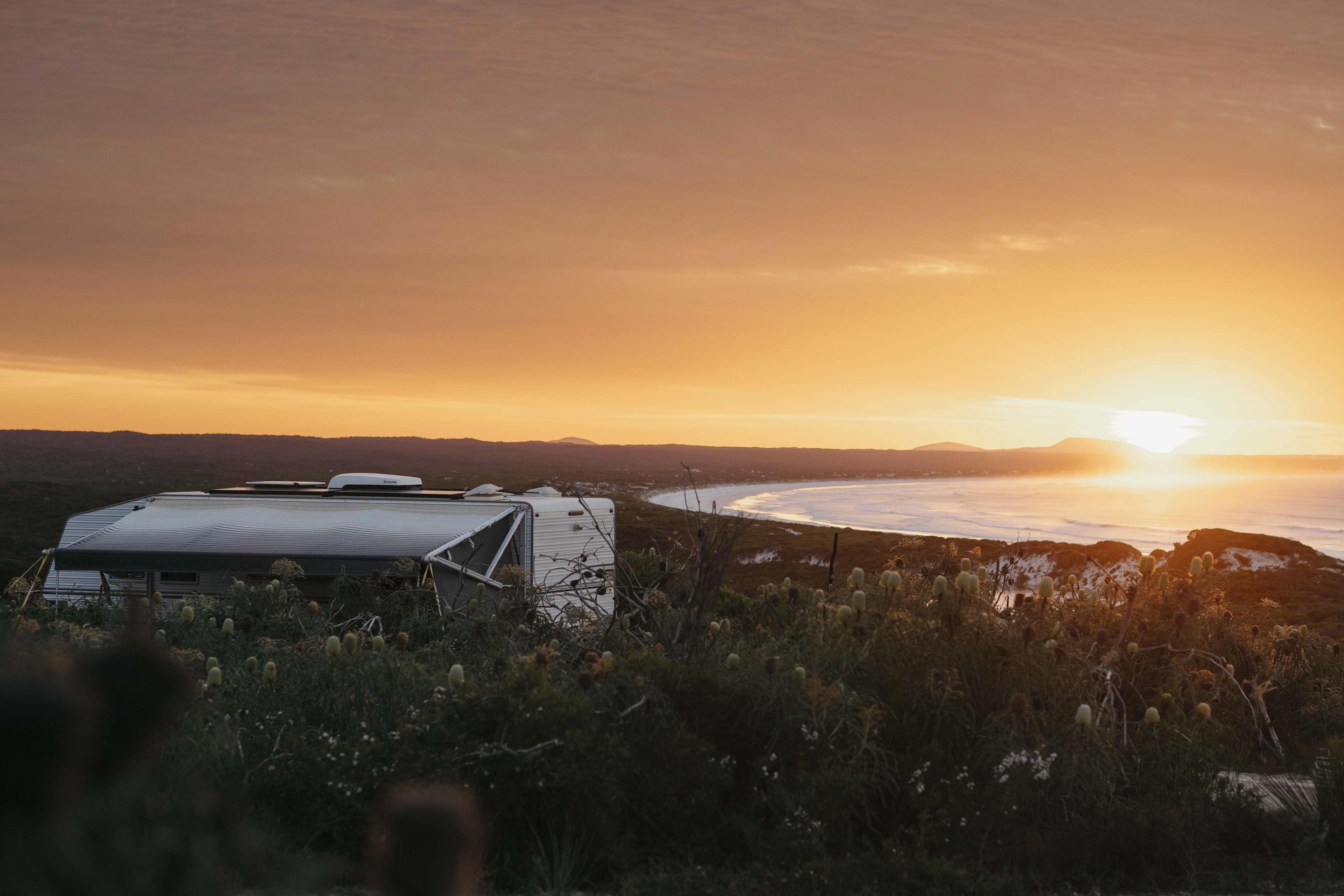 The top of a caravan is seen above low bushland as the sun sets over a beach.