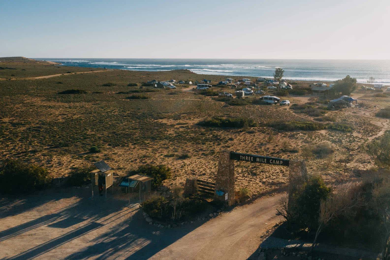 A road with a sign overhead that reads 'Three Mile Camp' leading to a campground with tents and cars, and ocean in the distance.