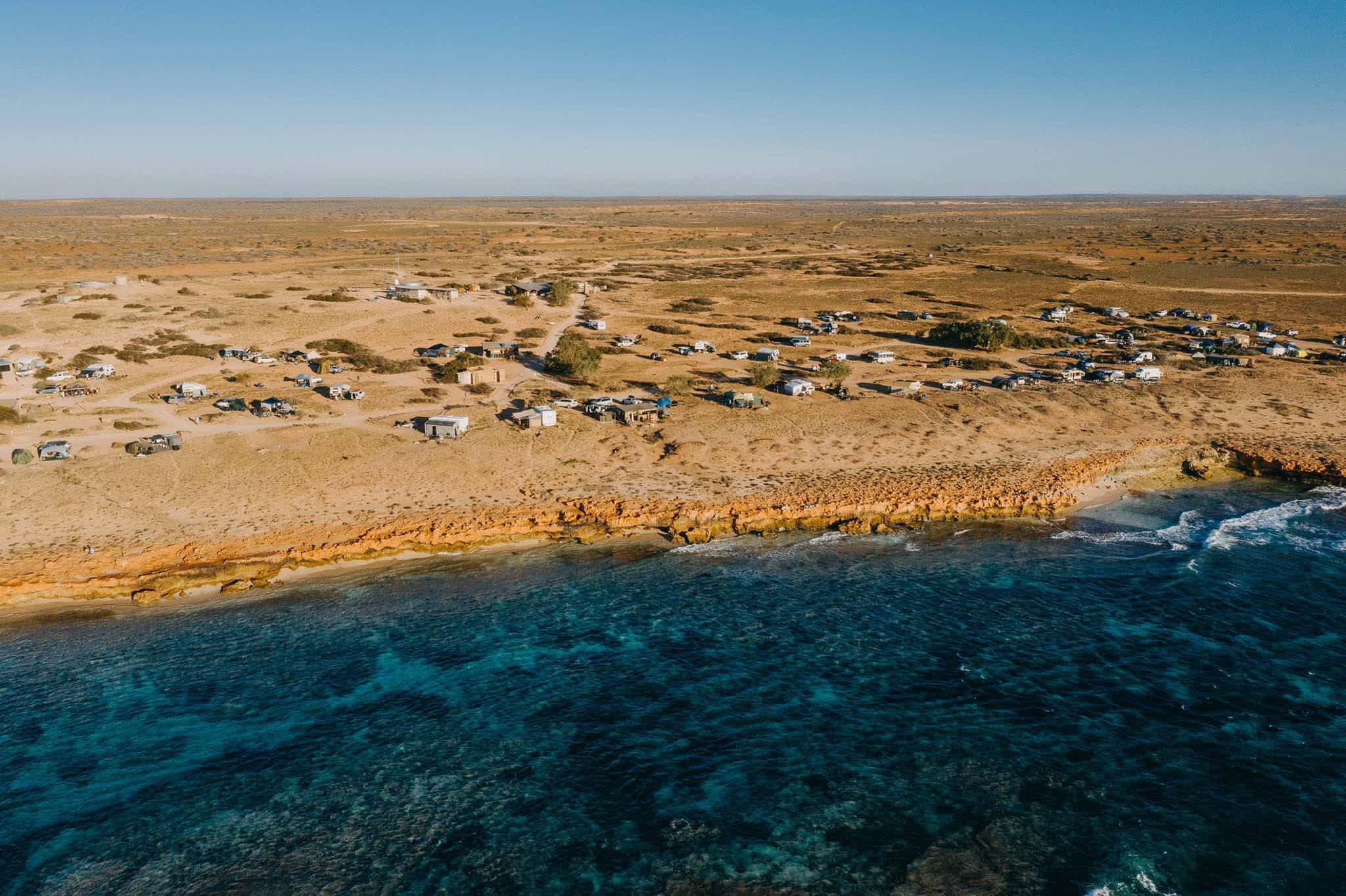 A drone shot of a campground set in a flat rocky landscape, with blue ocean and visible reef in the foreground.