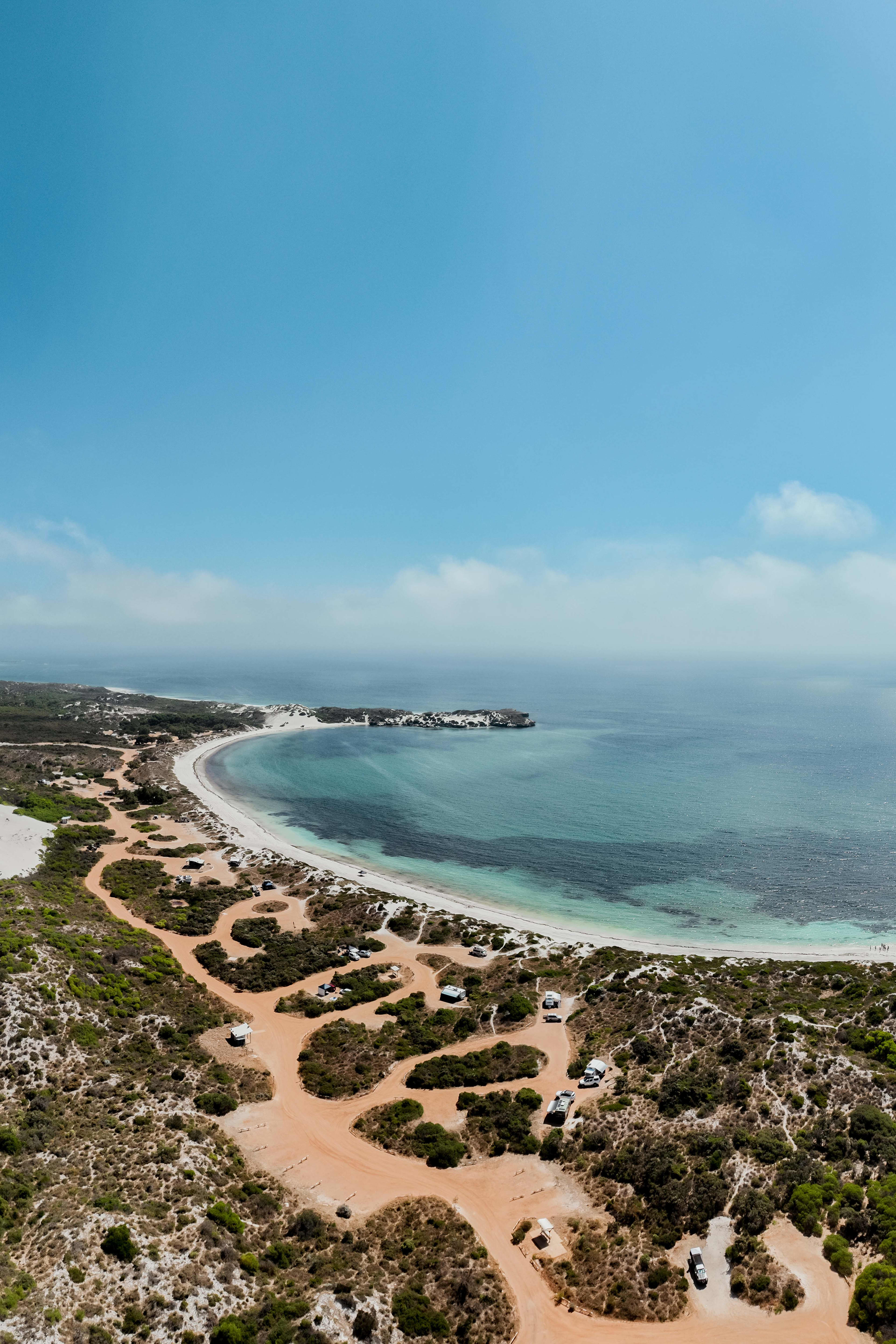 An aerial view of a campground, showing cleared sites and road in light sand/dirt between green coastal shrubs, with the bay in the background.