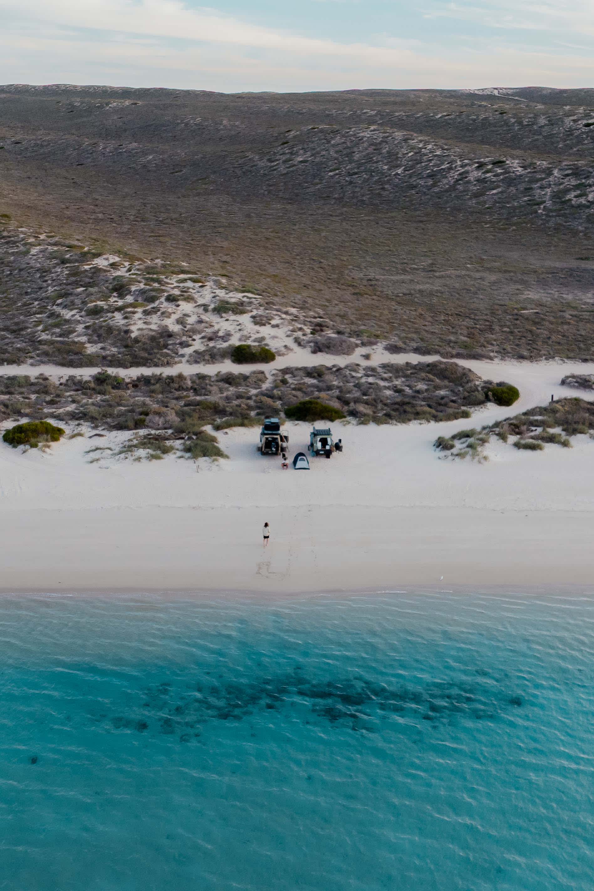 A drone shot showing blue ocean in the foreground, a vehicle and tent set-up on the white sandy beach, and a low green scrub-dominated landscape to the horizon.