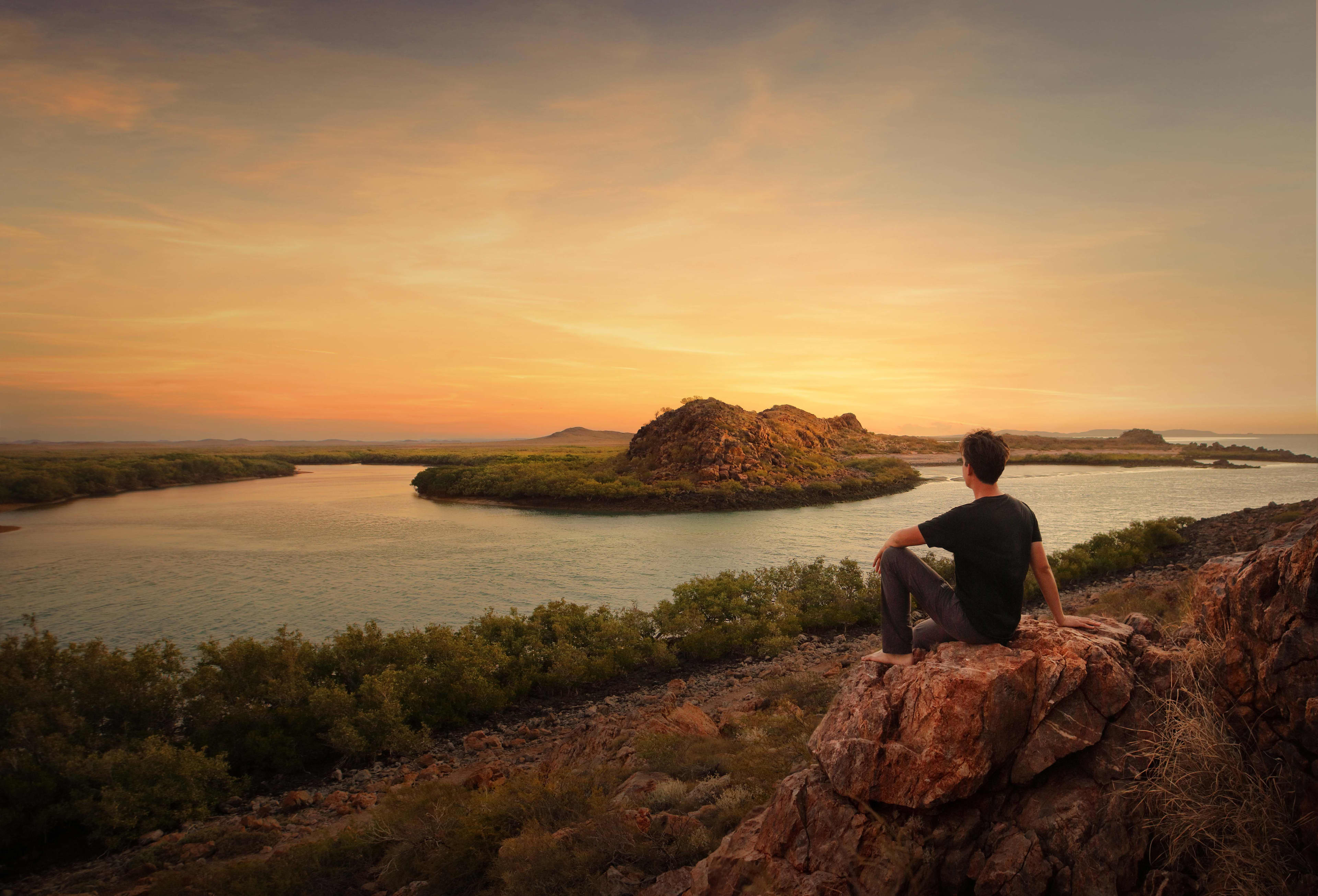 A person in a black shirt sits on a rock overlooking a landscape of a wide river with a rocky island in the middle.