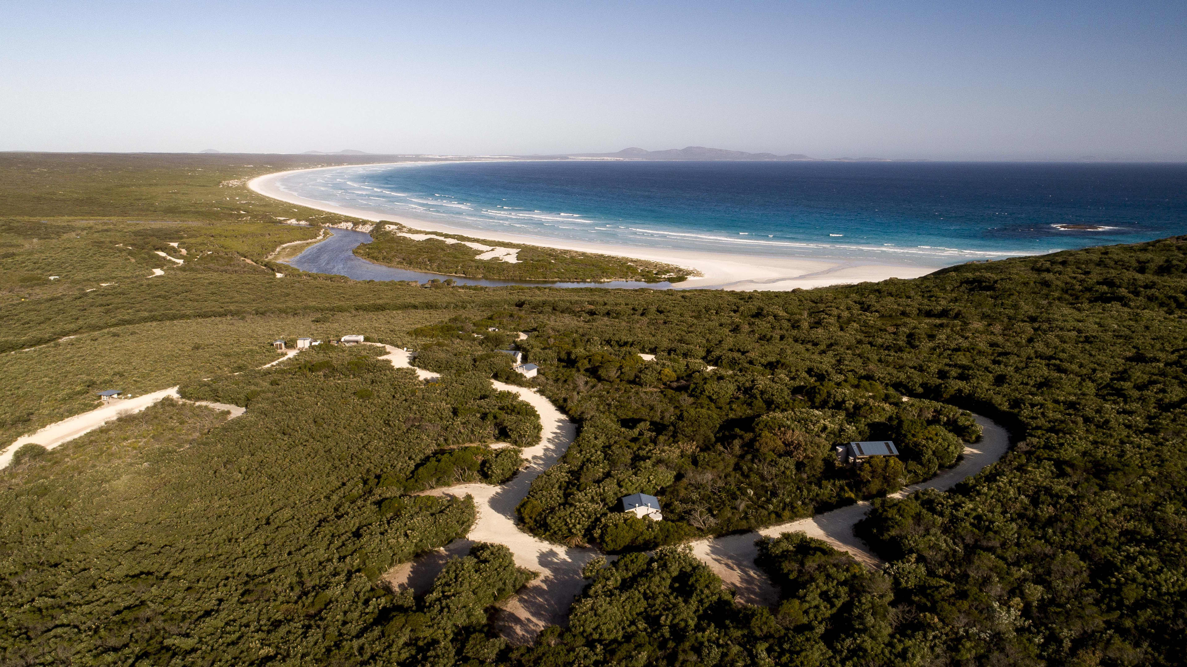 An aerial photo of a campground showing white sandy roads and sites among green bushland, with the beach and ocean in the background.