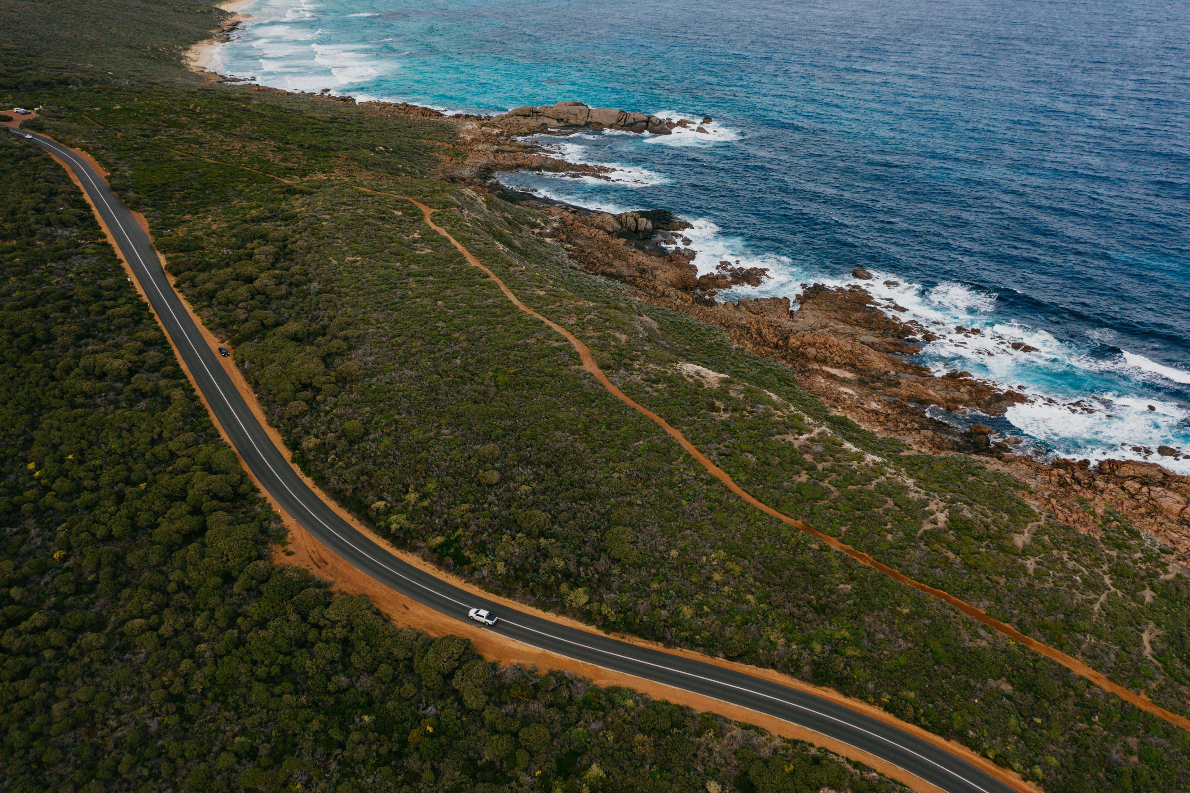 A white car appears as a small dot on a winding bitumen road through green bushland, with the rocky shore and ocean nearby.