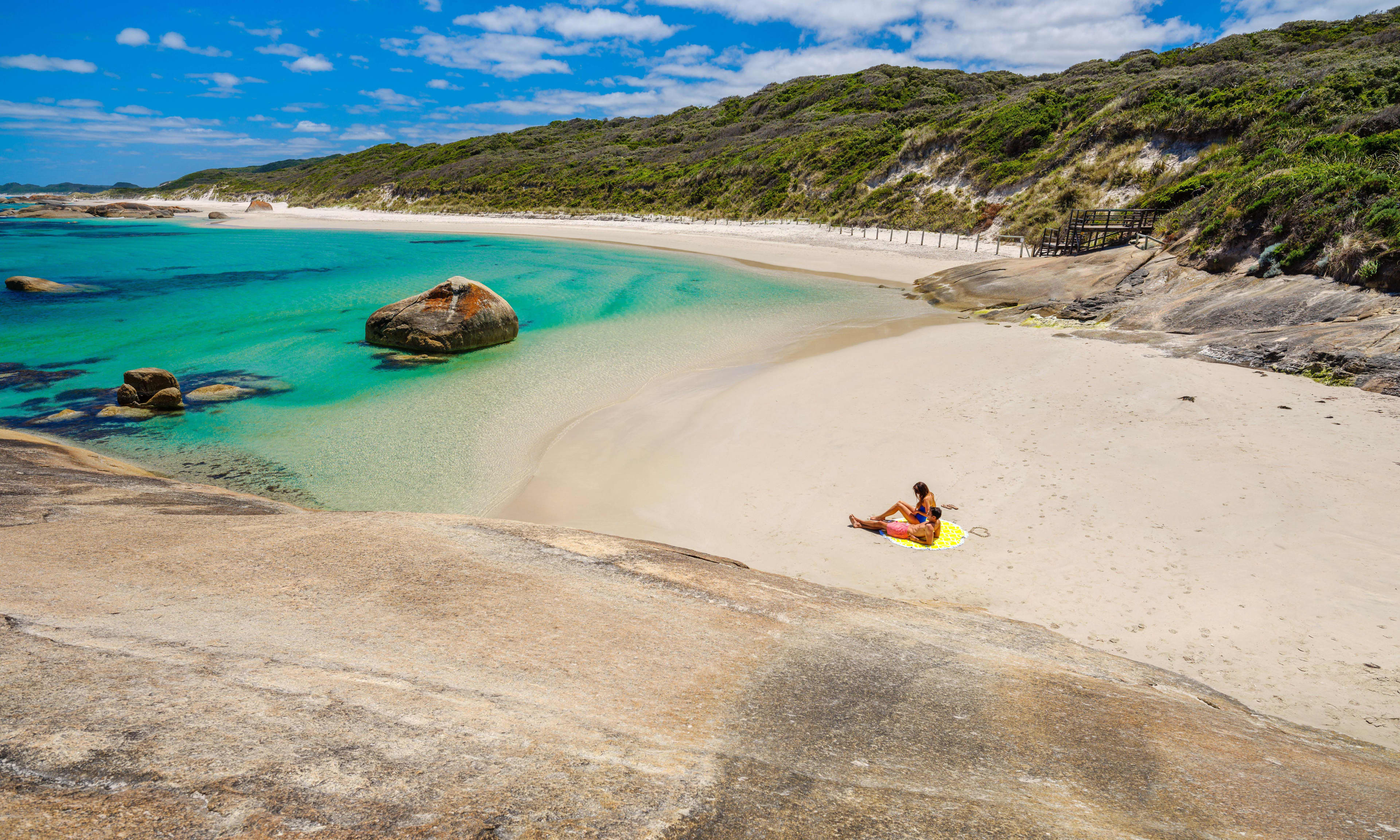 A person sits on a yellow beach towel on the sand, with blue ocean and rocky outcrops, with greenery-covered dunes.