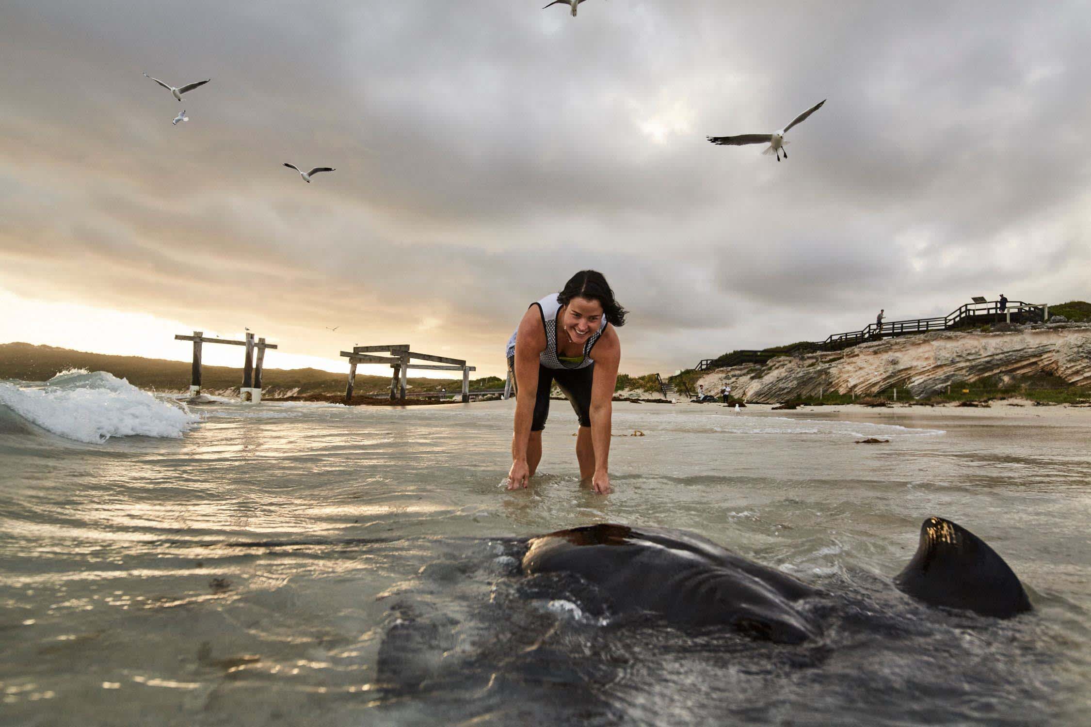 A woman bends to have her hands in the water, with a black stingray in the foreground and a cloudy sky with seagulls flying overhead.
