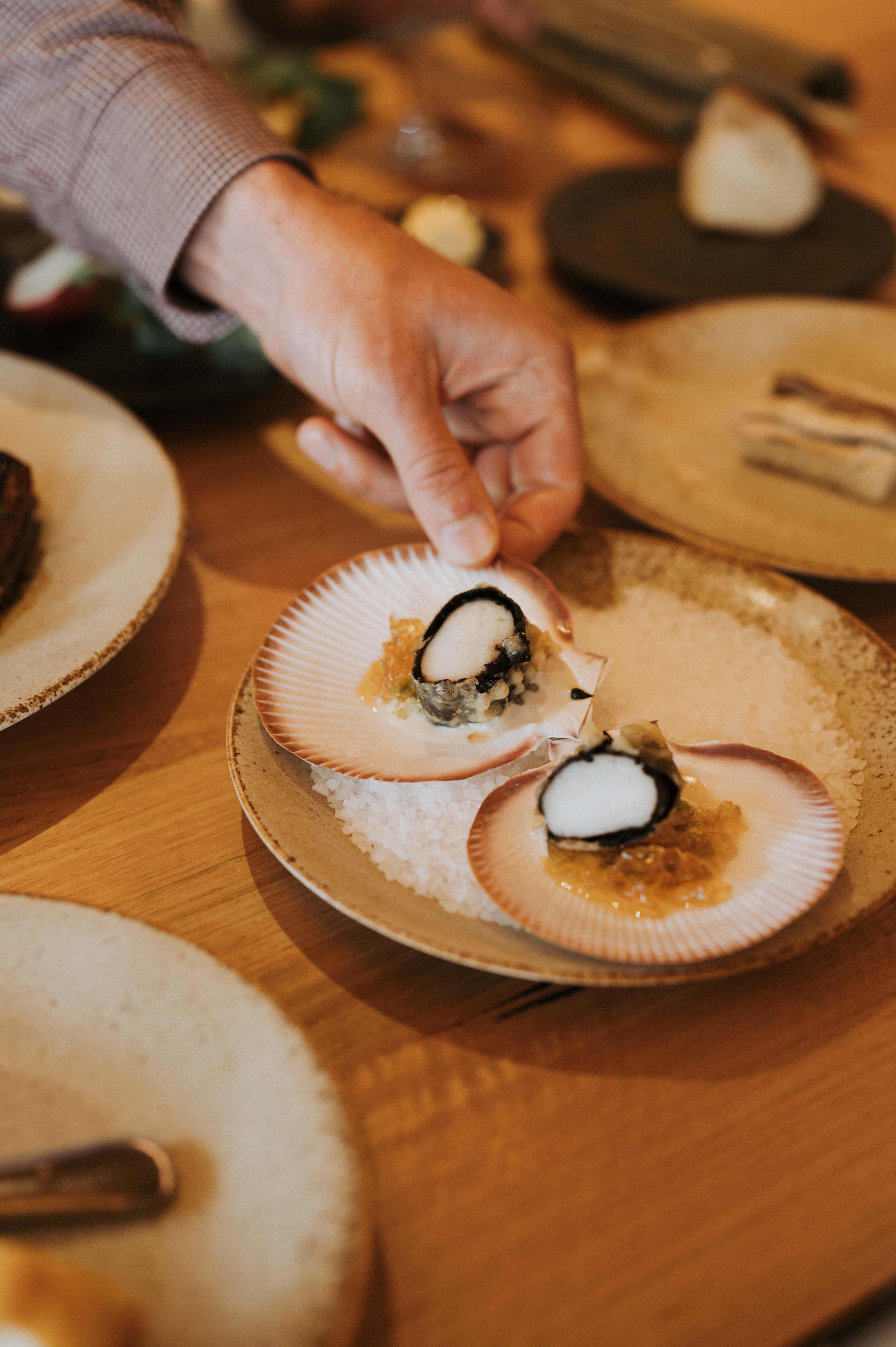 A hand reaches out to pick up a scallop from a plate with two on a restaurant table.