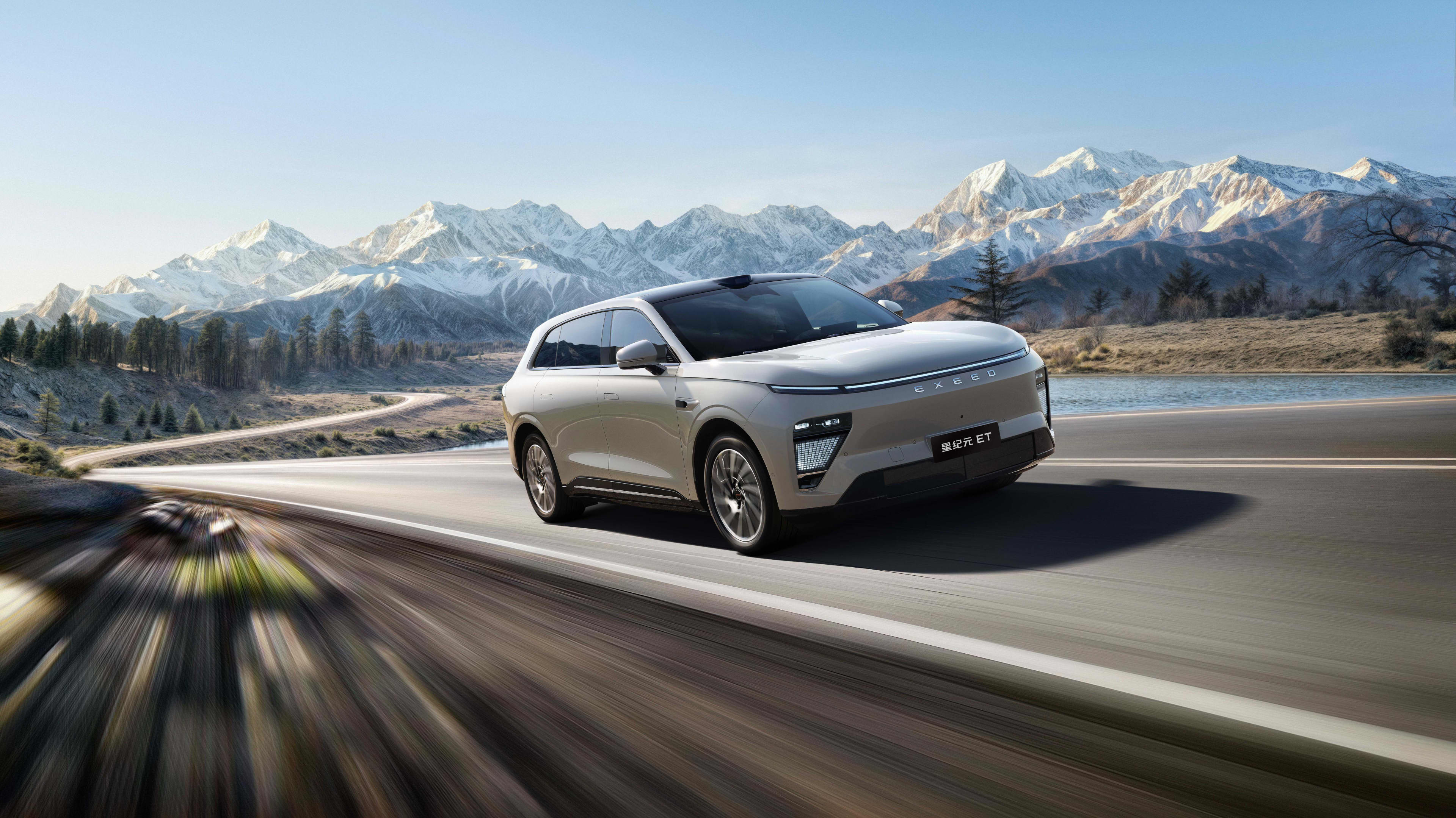 A pale coloured car driving along a winding bitumen road, with snow-capped mountains in the background.