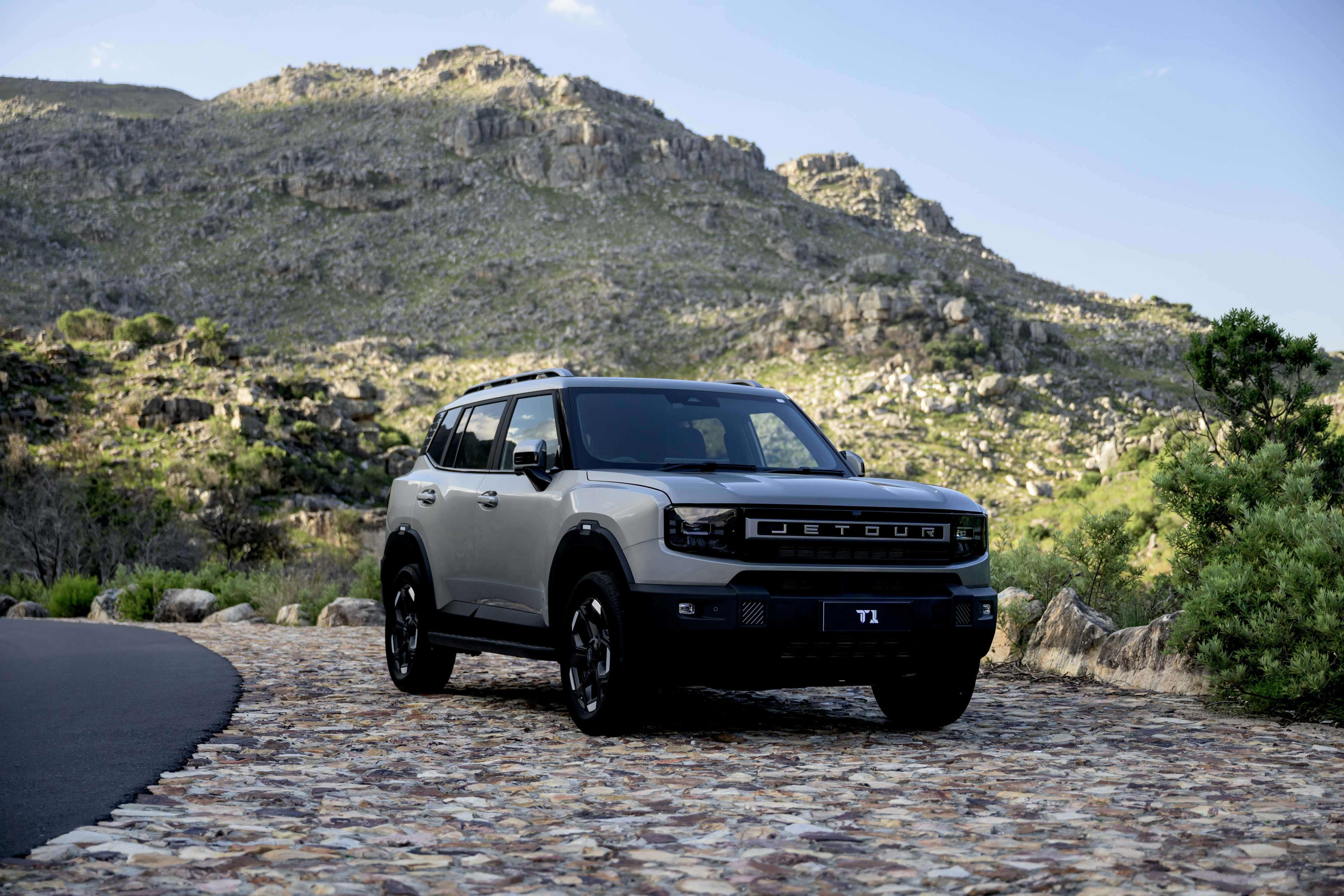 A light coloured SUV parked on a cobblestone road with a rocky, green hill in the background.
