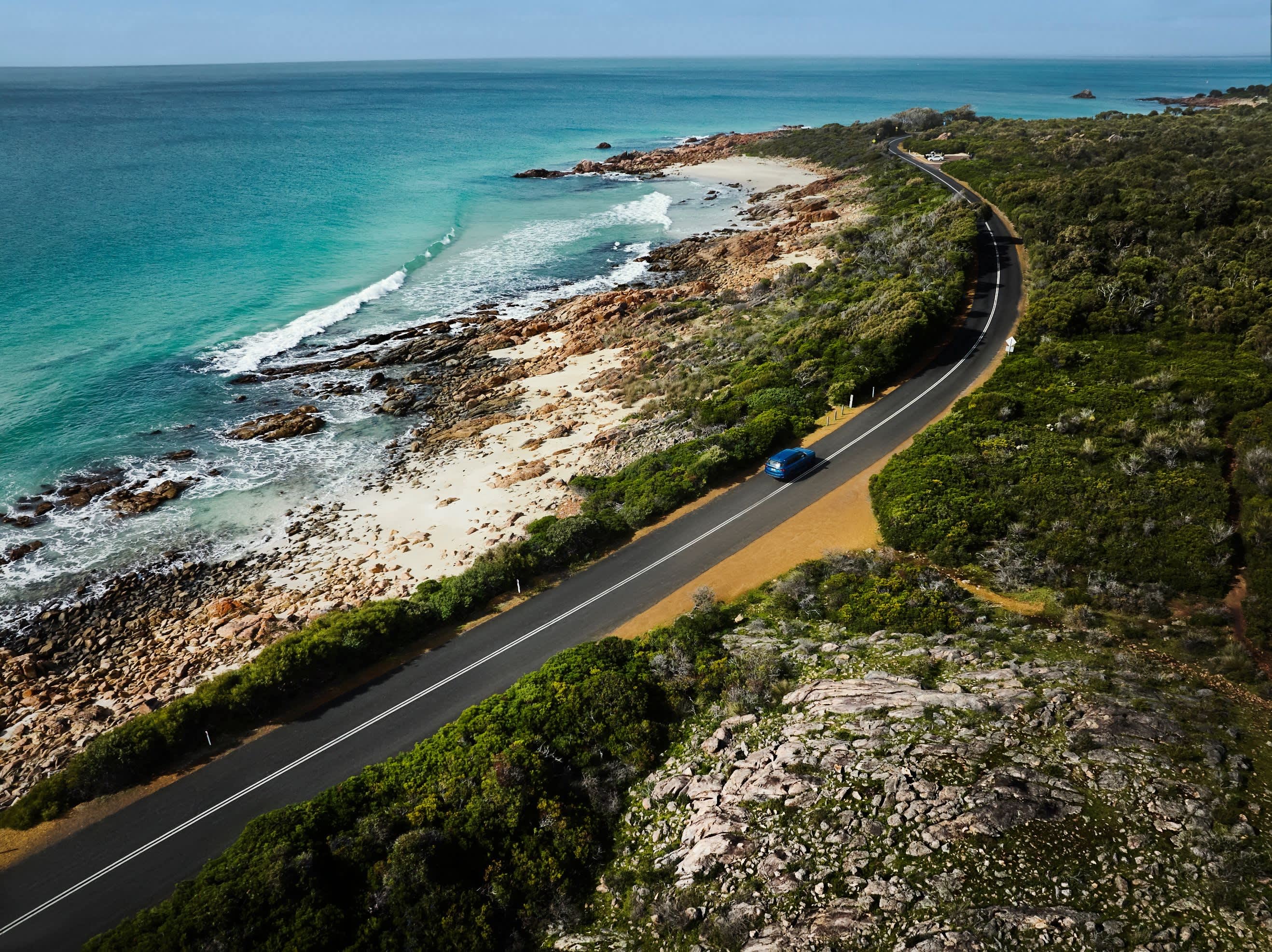 A blue car drives along a bitumen road, with bushland on one side and the ocean on the other.