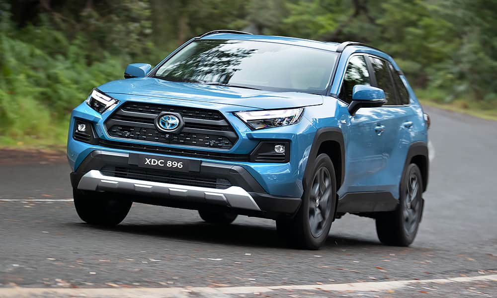 A light-blue Toyota RAV4 driving on a country road edged with green foliage