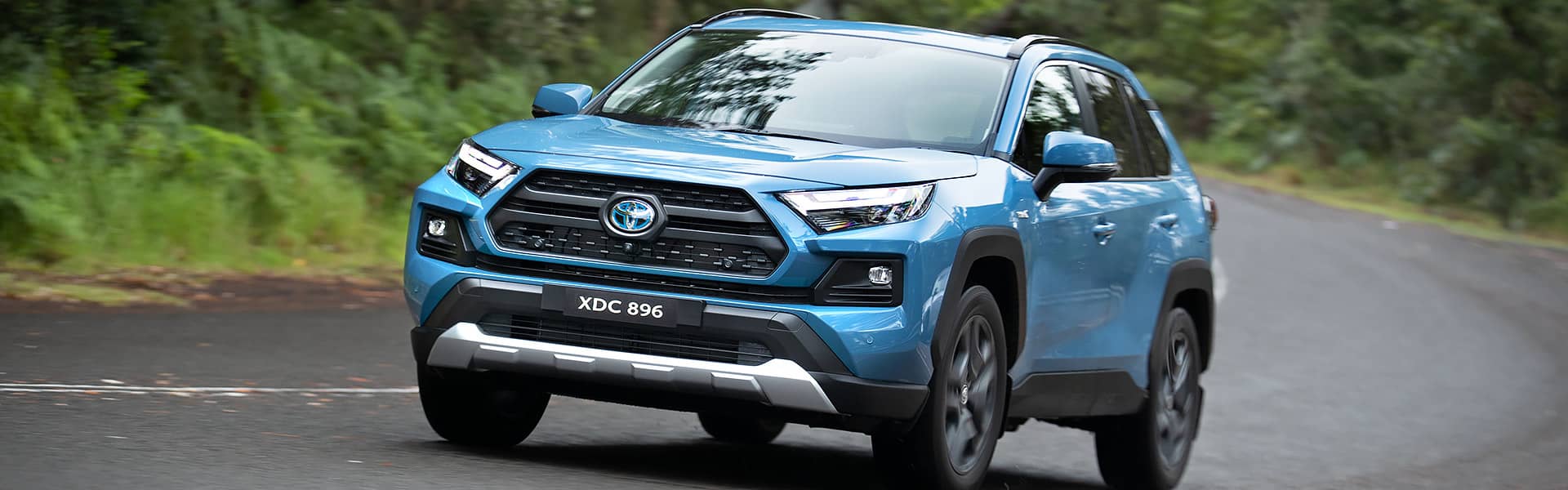 A light-blue Toyota RAV4 driving on a country road edged with green foliage