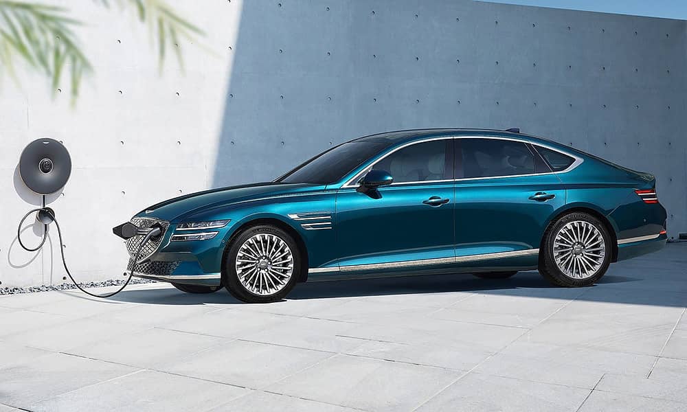 A blue Genesis G80 Electrified being charged with a white concrete wall in the background