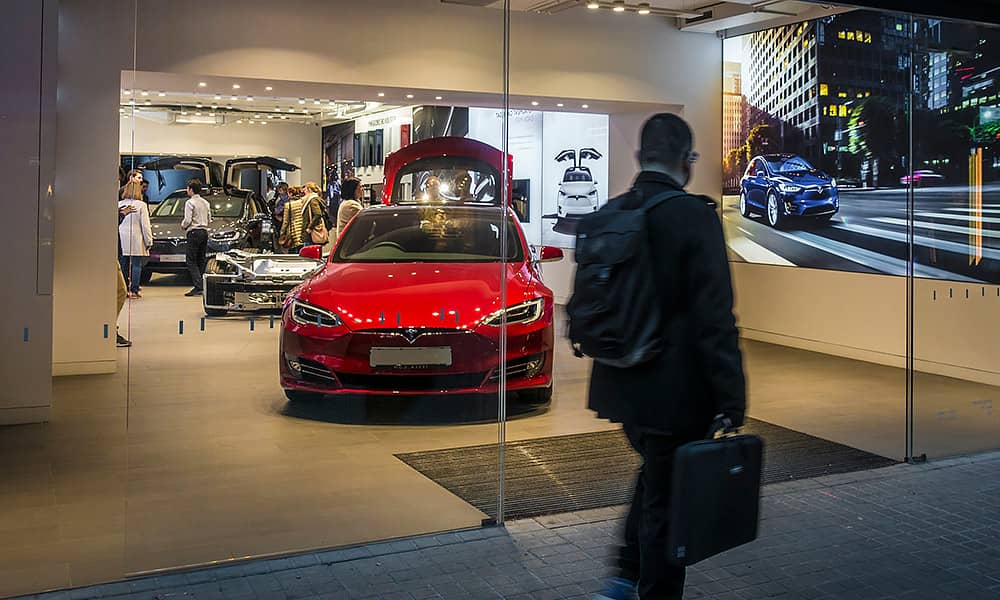 A man in a business suit  walking past a Tesla showroom