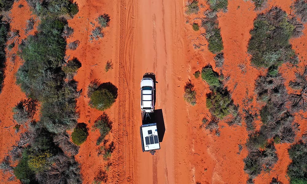 An aerial shot of a car and caravan travelling down a red dirt road